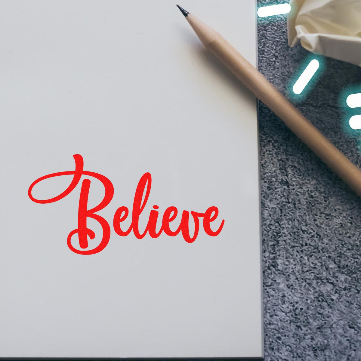 Red Believe stamp seal on a white notebook beside a tan pencil, on a gray textured surface.