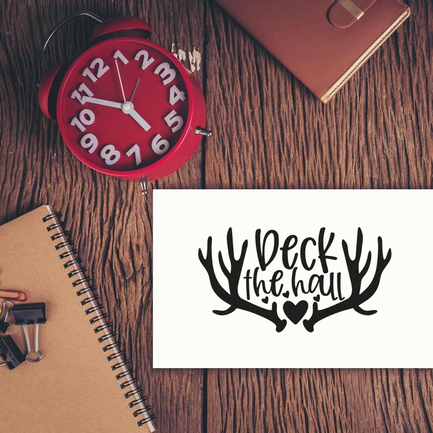 Black antler holiday stamp and seal design on white card, beside a red alarm clock on a wood desk.