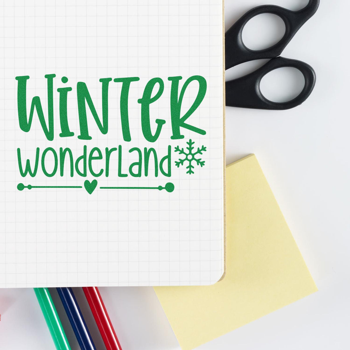 Green Winter Wonderland stamp and seal design on graph notebook, with black scissors, yellow sticky note, and colored markers.