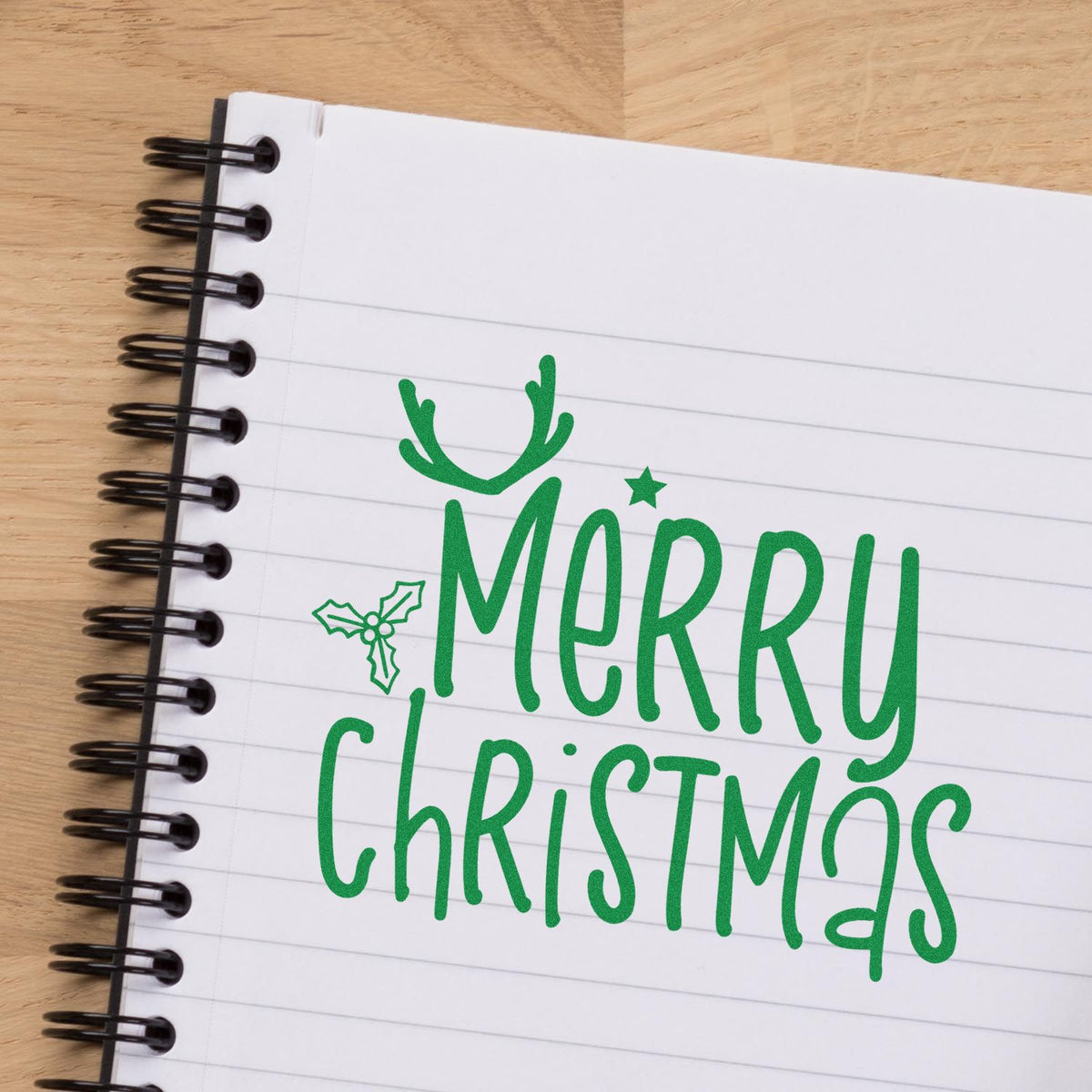 Green Merry Christmas stamp with holly and antlers on a white spiral notebook over a wood tabletop.