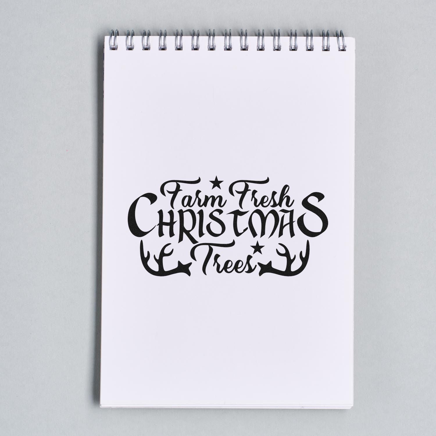 Black Farm Fresh Christmas Trees stamp design on a white spiral notebook, with antlers and stars for holiday seal products.