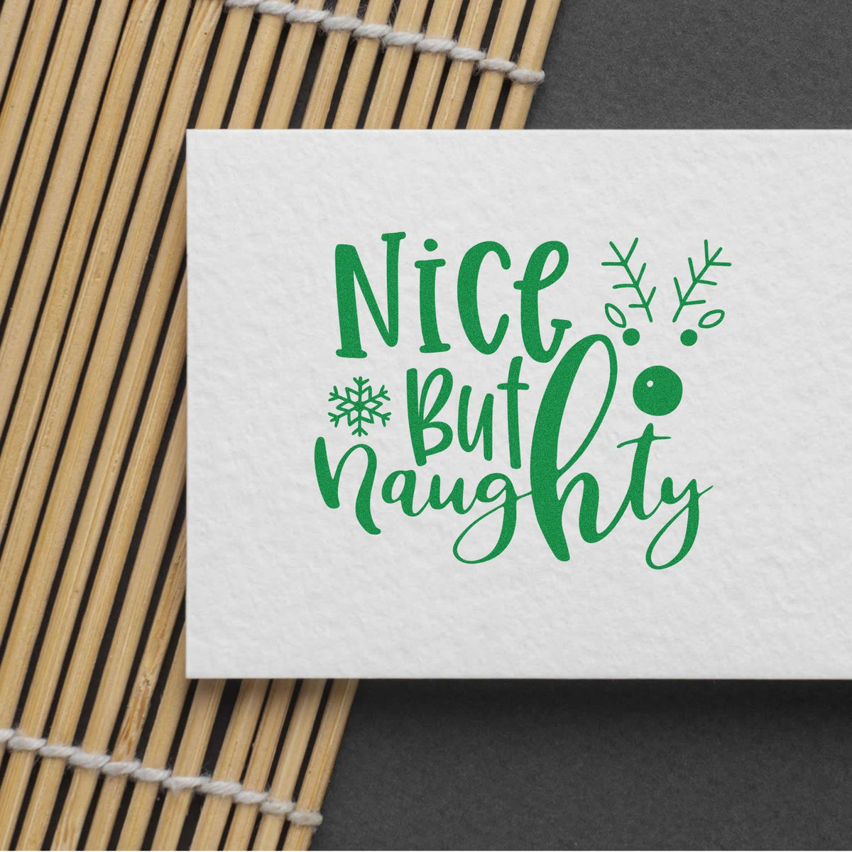 Green Nice But Naughty holiday stamp and seal design on white card with snowflakes, on bamboo mat.
