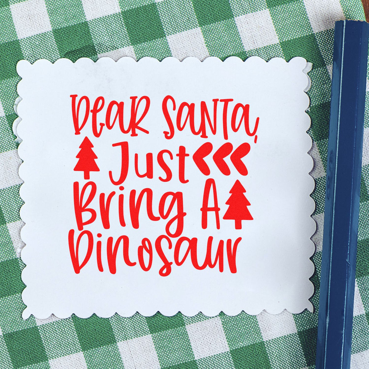 White scalloped stamp/seal with red Dear Santa, Just Bring a Dinosaur text and tree icons on green plaid, blue tool at right.