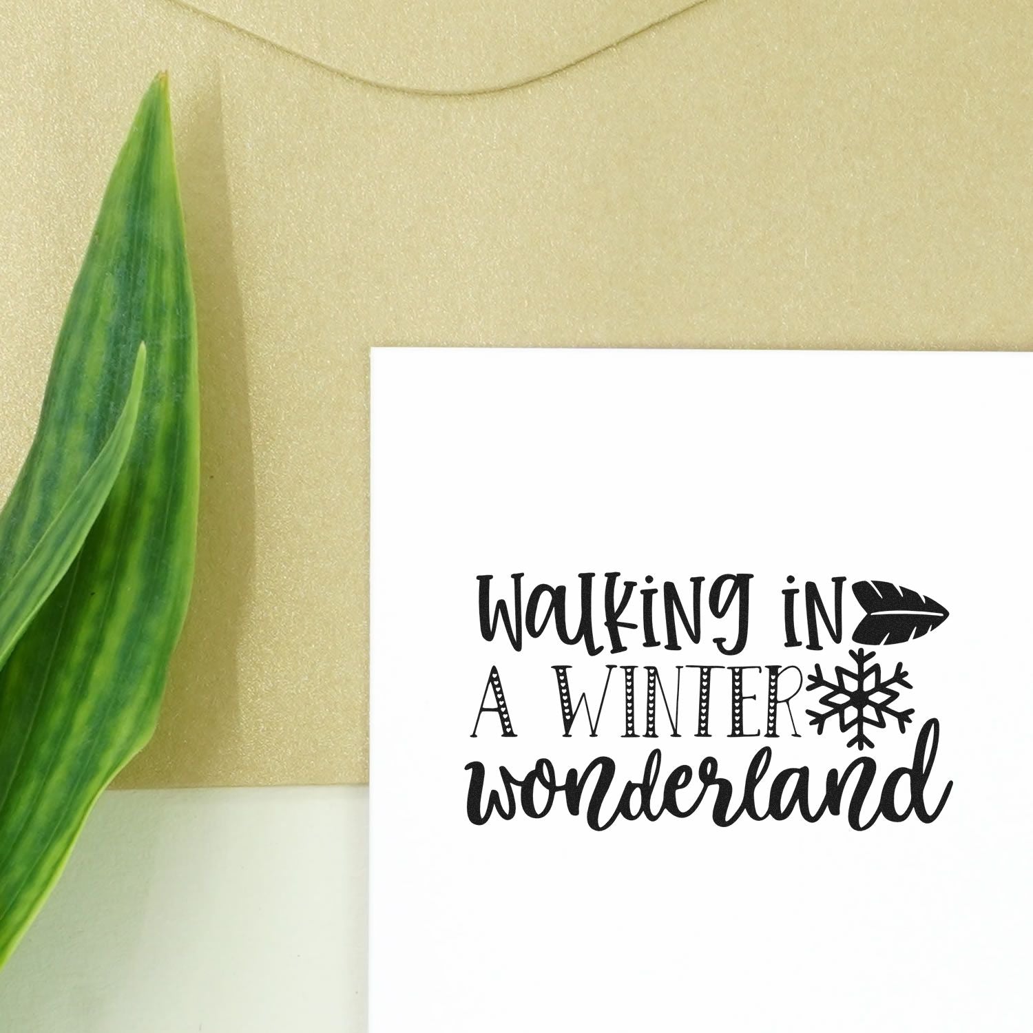 Black winter wonderland stamp and seal on white card with feather and snowflake accents, beside a green leaf on beige background