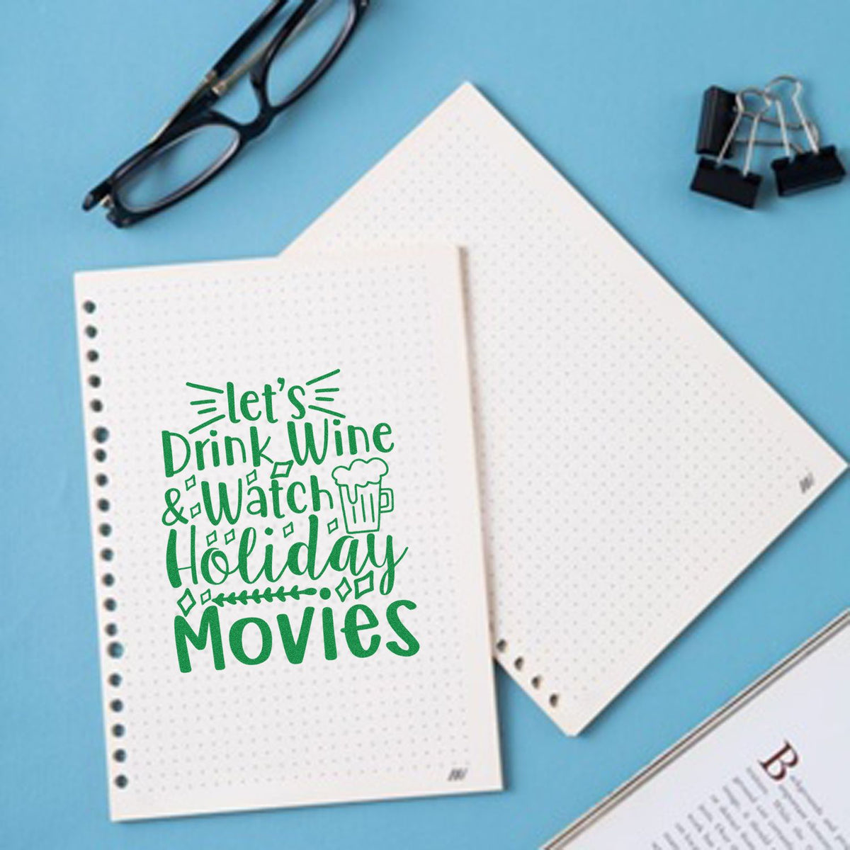 Green holiday movie stamp/seal on white dotted notebook pages, with black glasses and binder clips on a blue background.