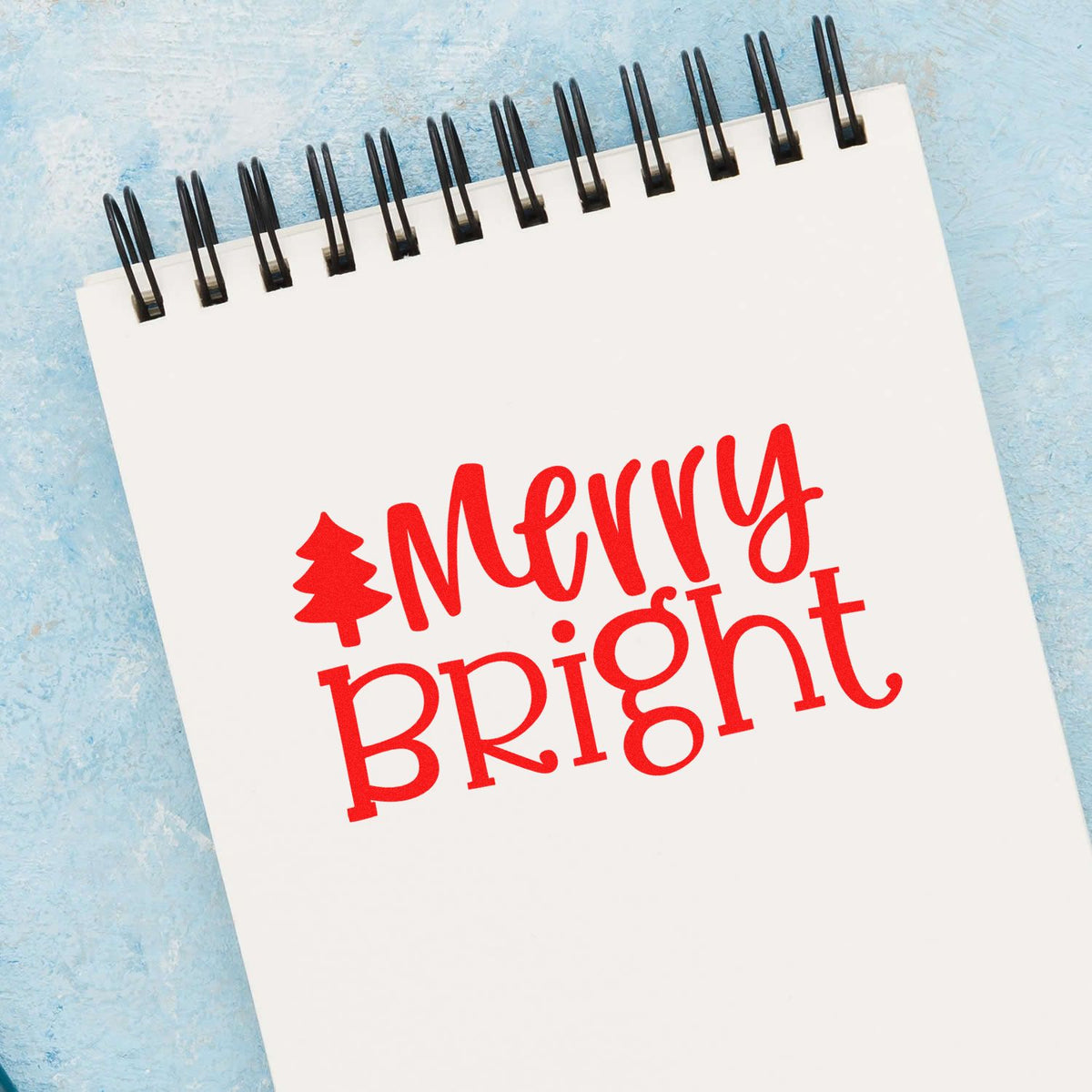 Red Merry Bright holiday stamp and seal design with tree icon on a white spiral notebook, on a light blue background.