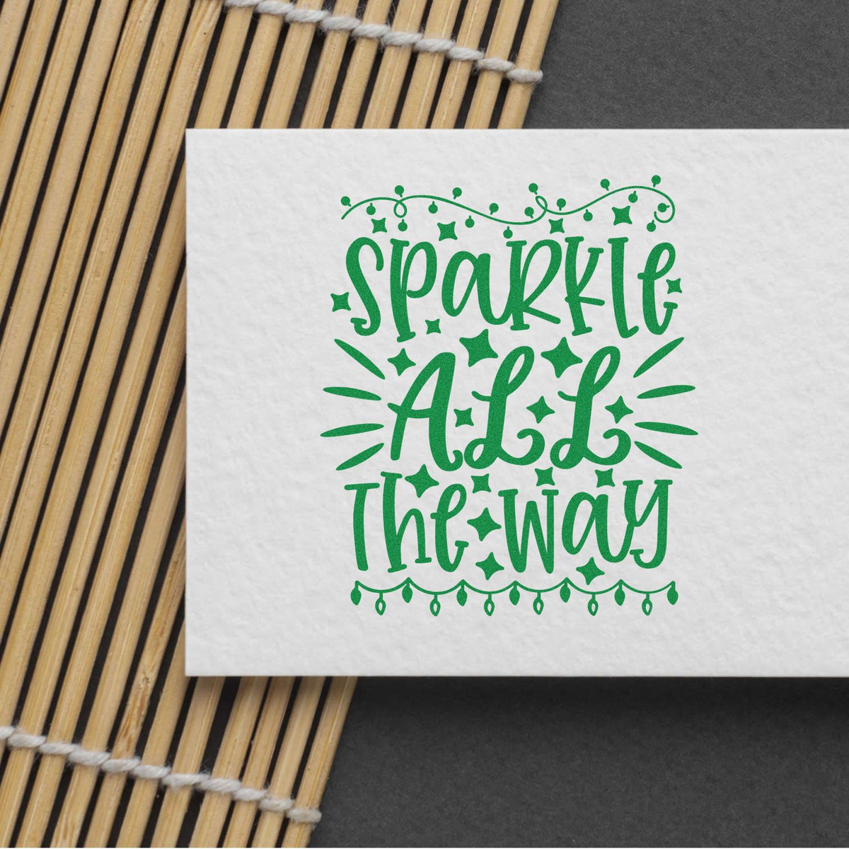 Green decorative stamp seal reading Sparkle Add The Way on white paper, with star and string accents beside bamboo mat.