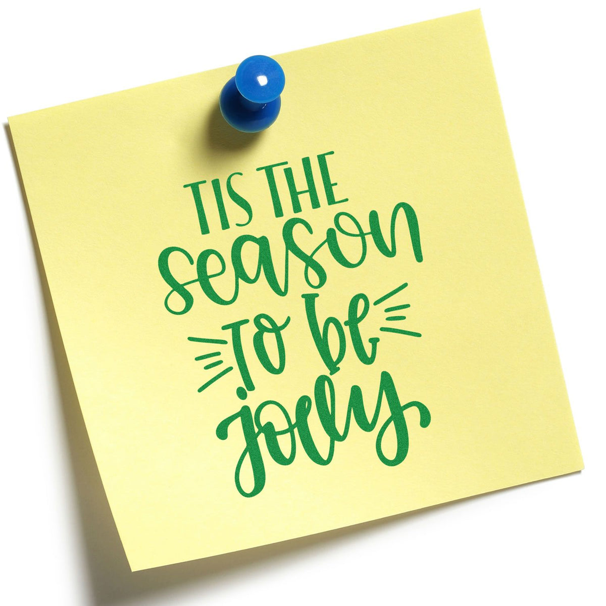 Yellow sticky note with blue pushpin and green holiday stamp-seal reading Tis the season to be jolly