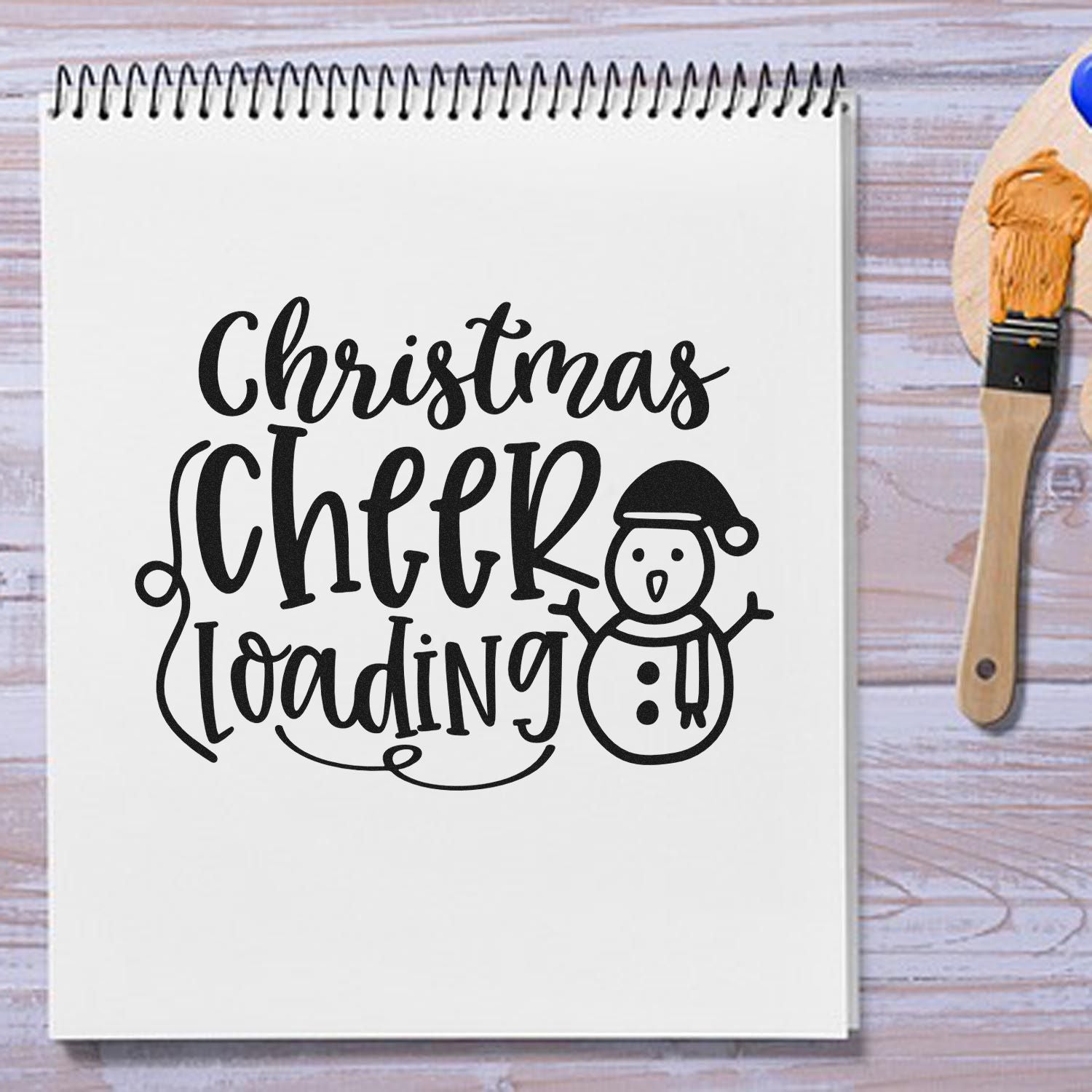 Black Christmas Cheer Loading stamp design with snowman on white notebook page, beside a wooden seal stamp with orange ink.