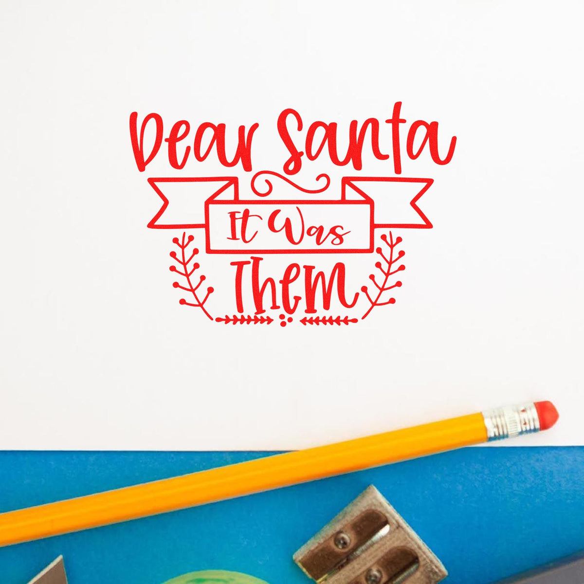 Red Christmas stamp and seal design reading Dear Santa It Was Them on white paper with pencil and blue background.