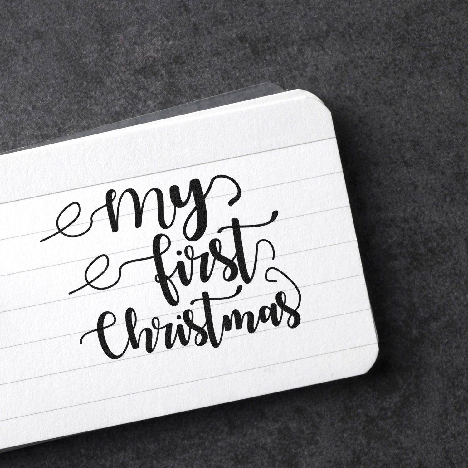 Black stamp and seal design reading "my first Christmas" on white lined paper against a dark gray textured background.