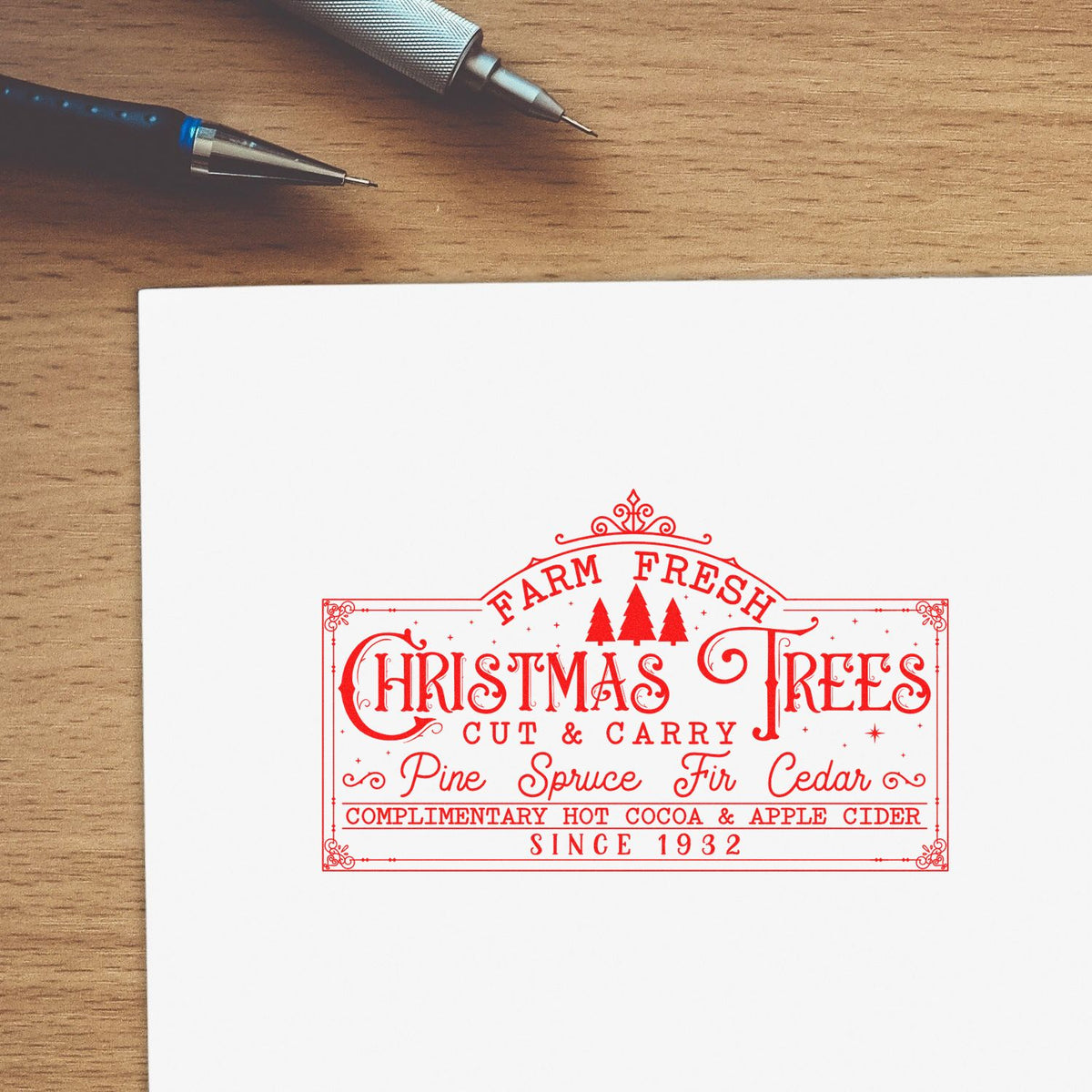 Red holiday stamp and seal design on white paper, featuring Christmas trees and festive text, beside two pens on a wood desk.