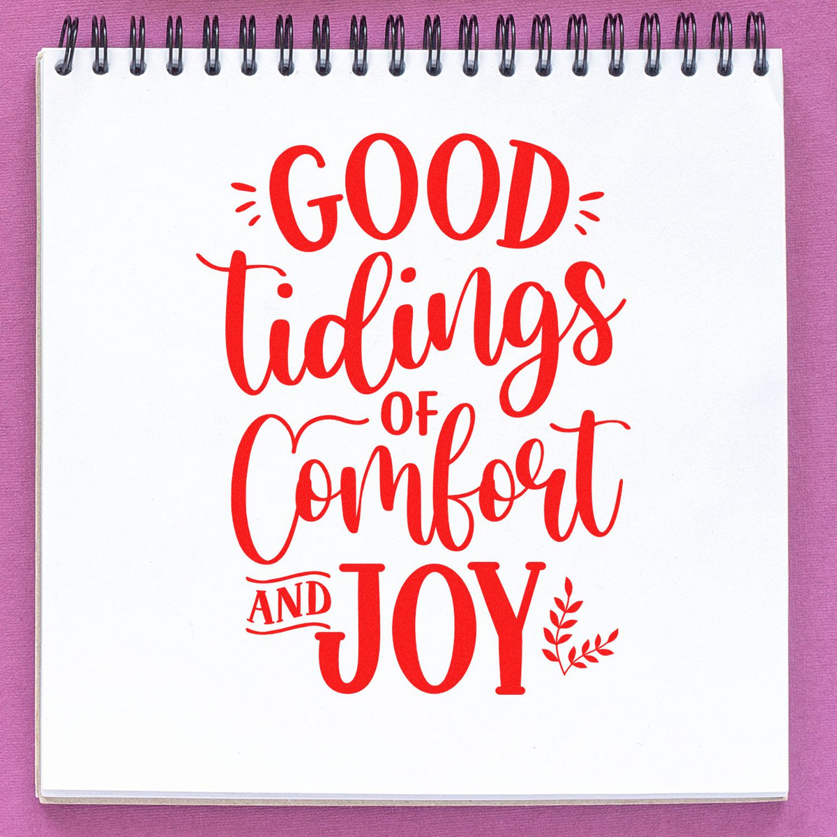 Red script stamp reading Good Tidings of Comfort and Joy on a white spiral notebook against a purple background.