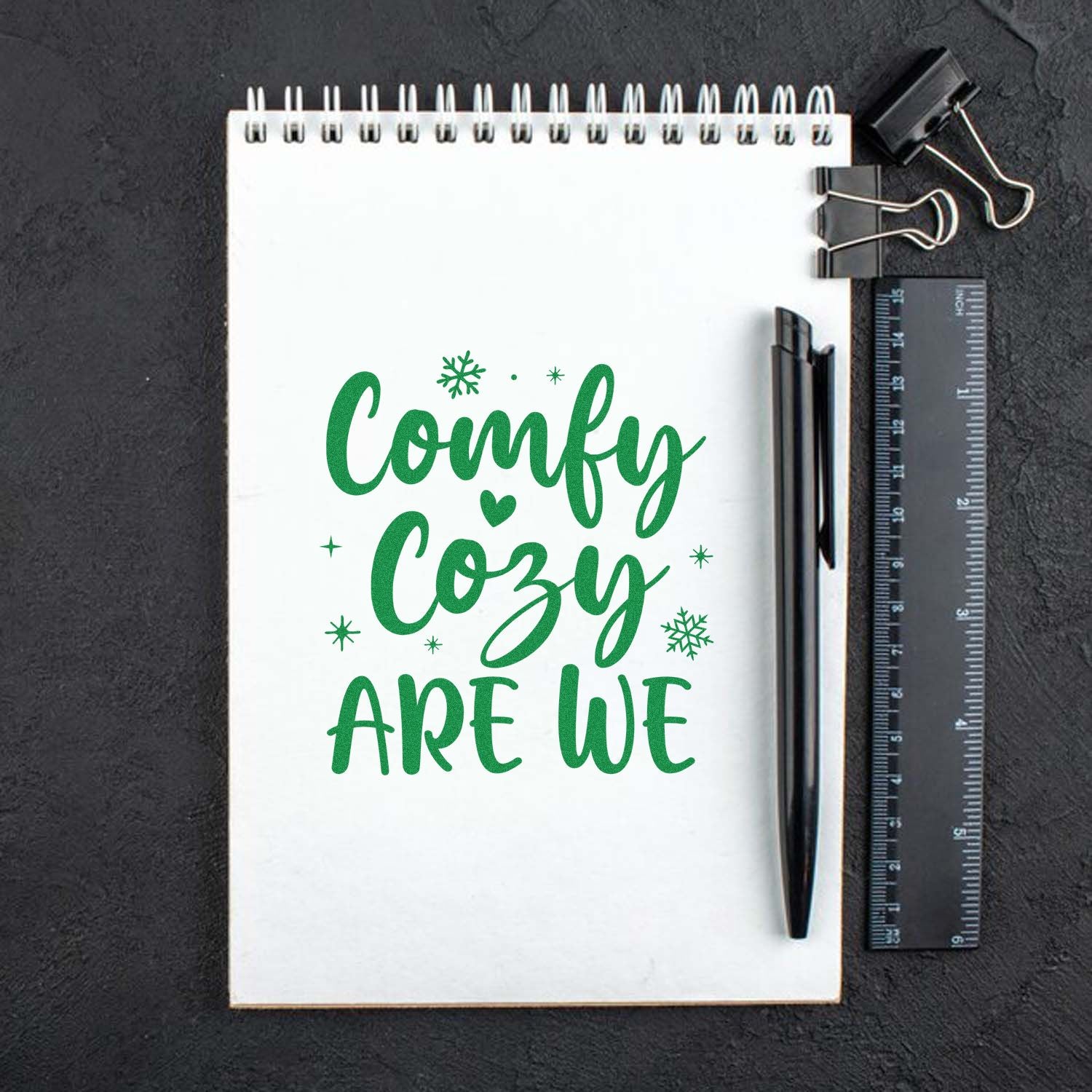 Green Comfy Cozy Are We stamp design on a white spiral notebook, with snowflakes and heart, beside a black pen and ruler.