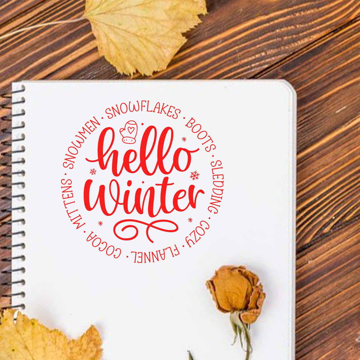 White spiral notebook with red hello winter stamp seal, circular winter words, on a wood table with dried leaves.