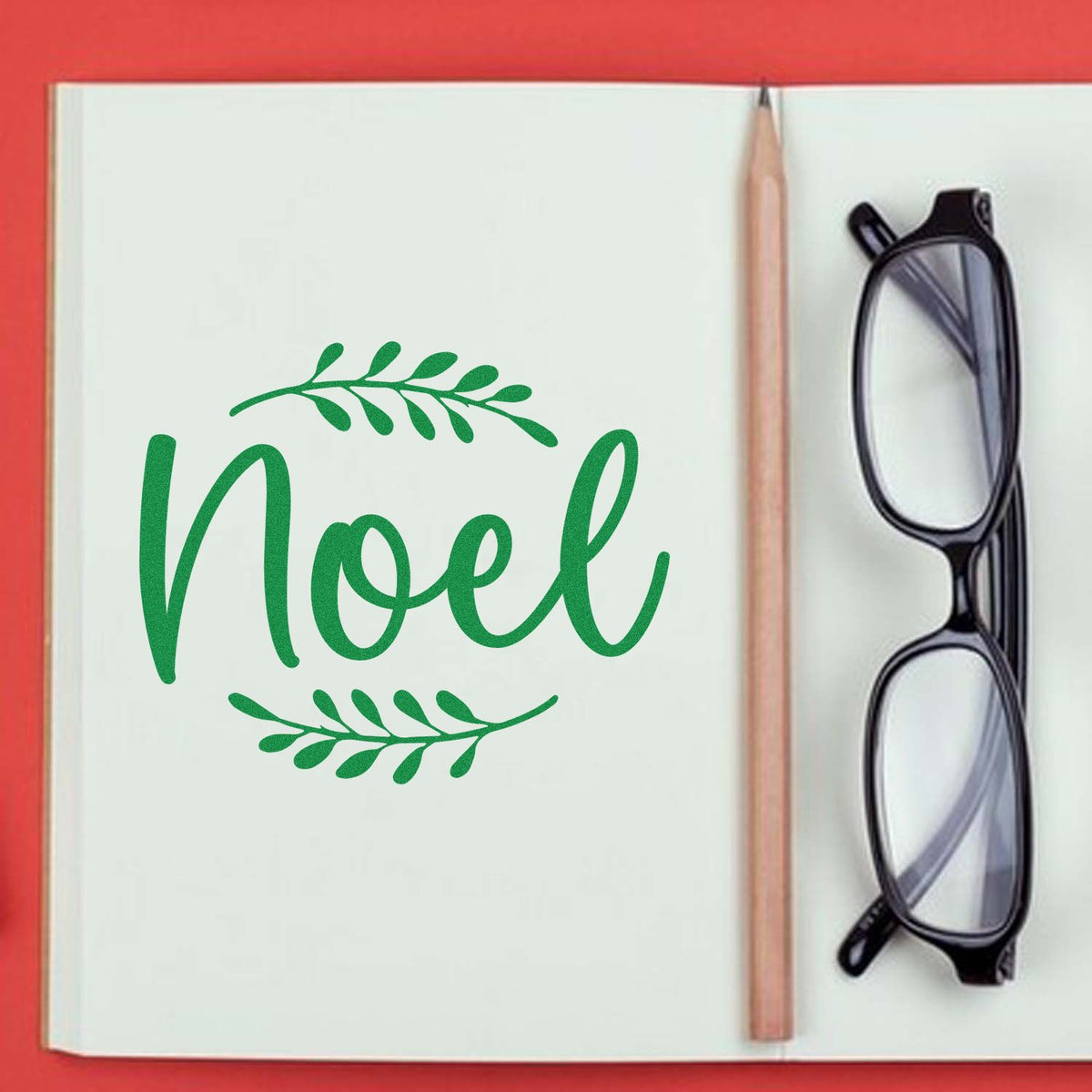 Green Noel stamp and seal with leafy wreath on white page, beside a pencil and black eyeglasses on a red background.