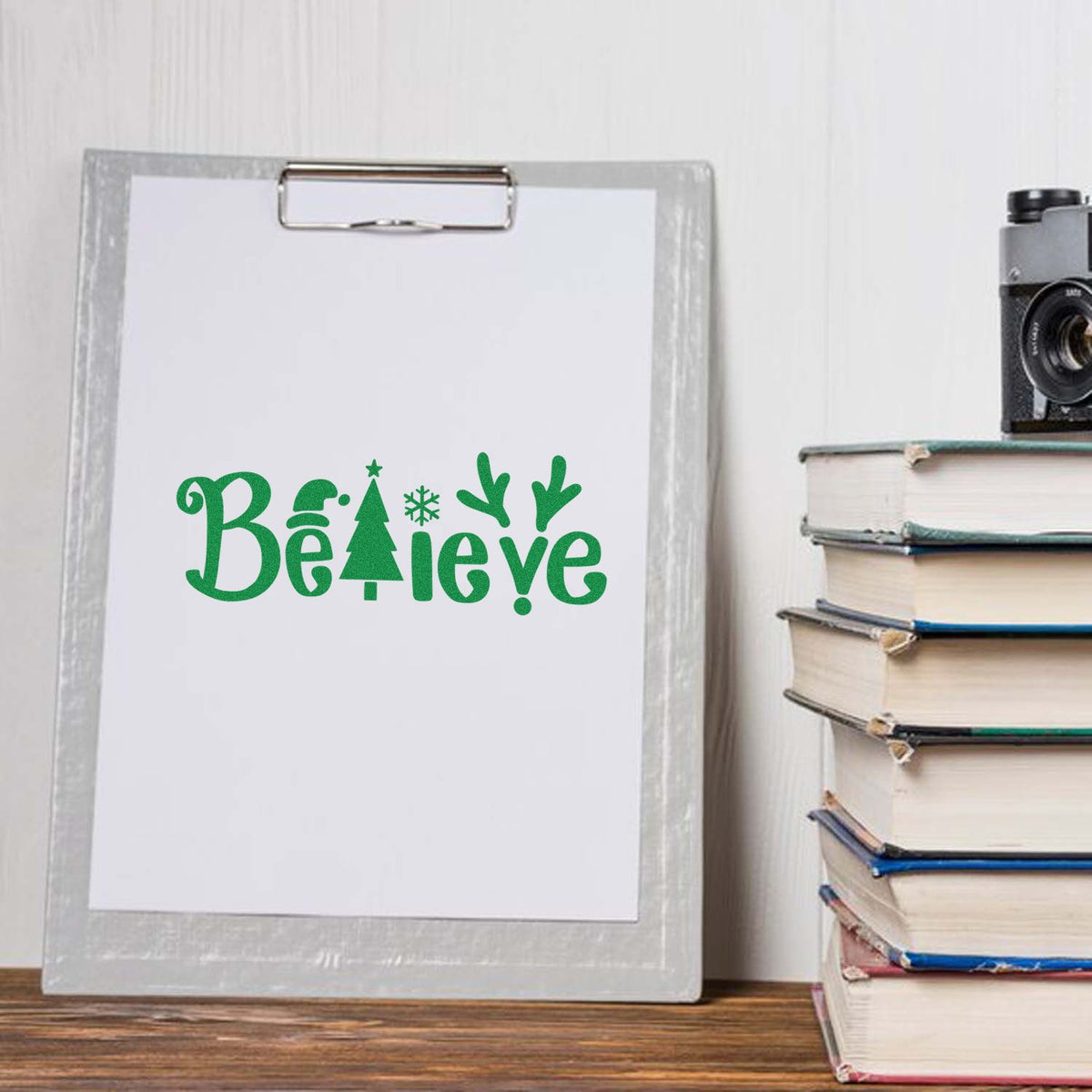 Green holiday stamp and seal design on a clipboard, with Believe text, tree, snowflake and antlers beside stacked books.