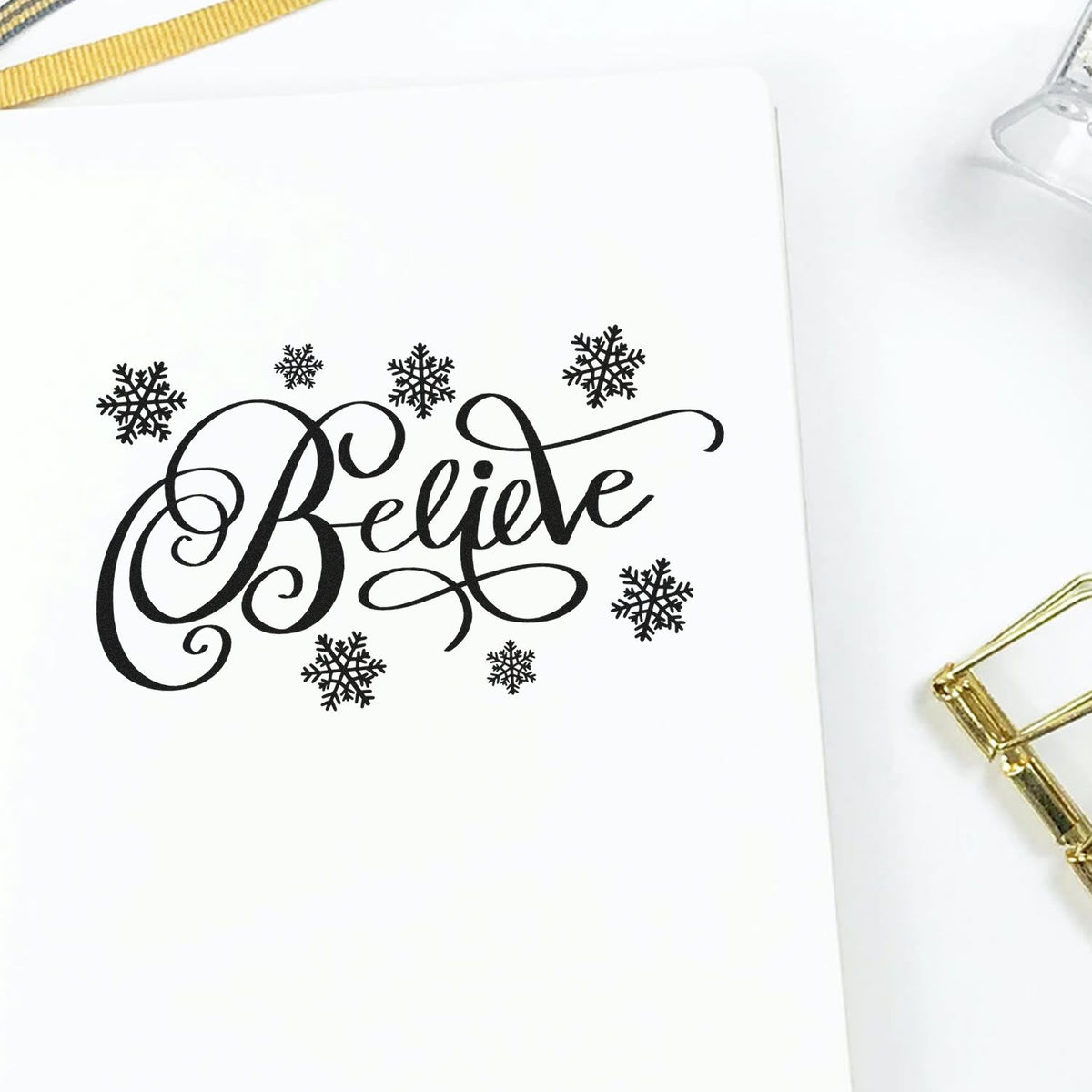Black Believe stamp and seal design with snowflakes on white paper, accented by gold ribbon and clip.