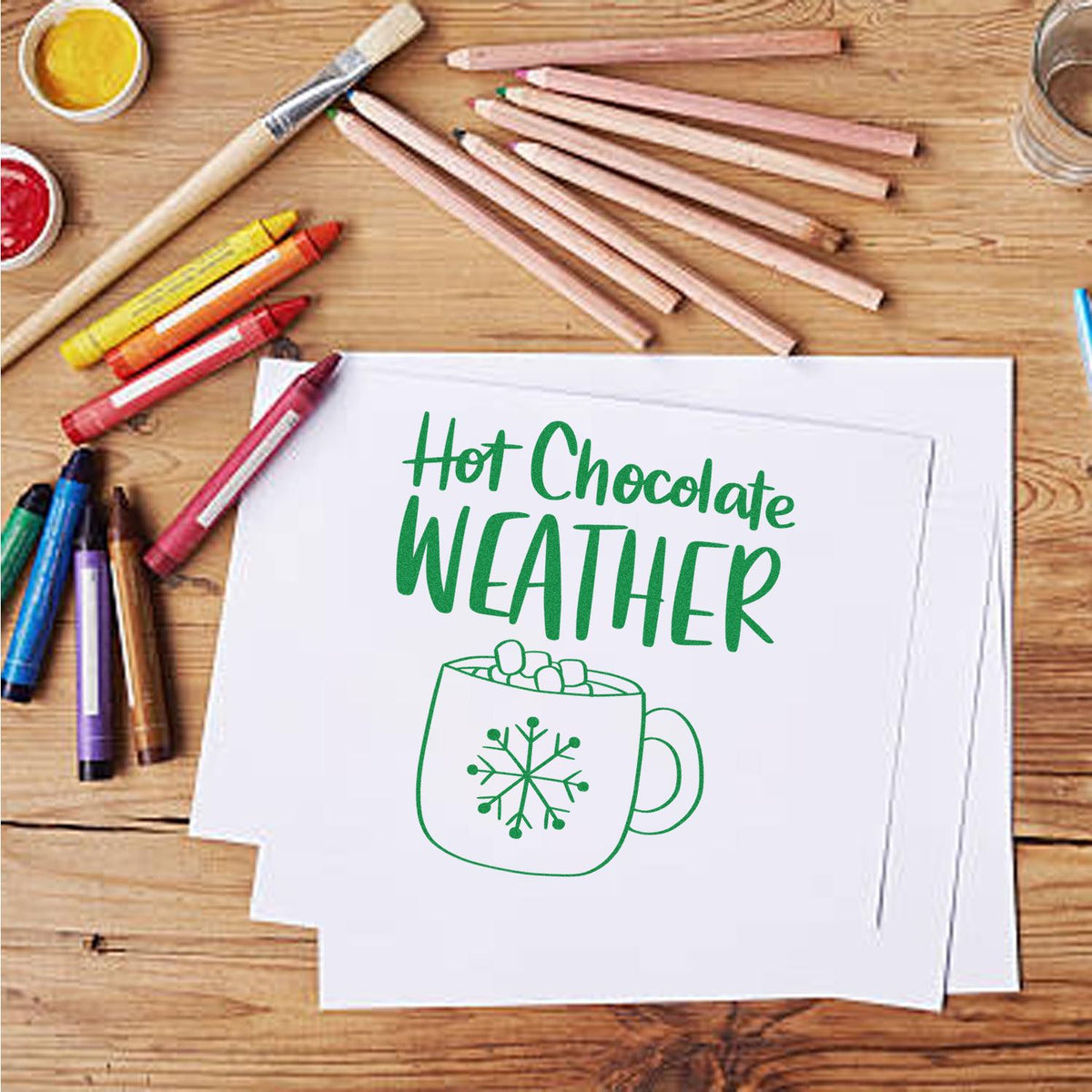 Green hot chocolate weather stamp and seal design with mug snowflake on white paper, surrounded by colored pencils.