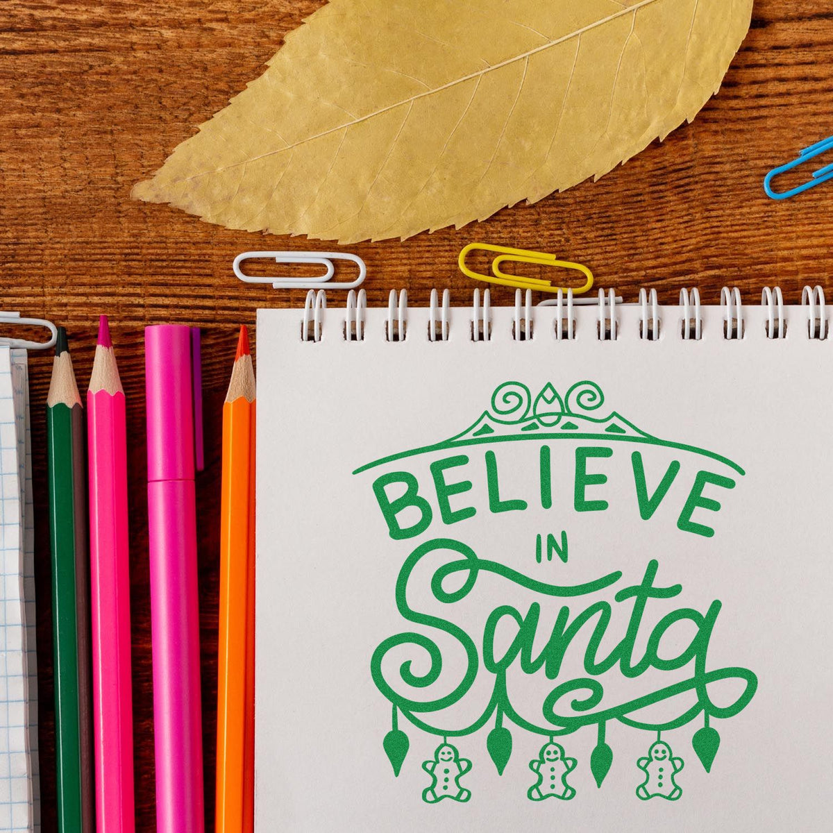 Green Believe in Santa stamp and seal design on white spiral notebook, with colored pencils and paper clips.