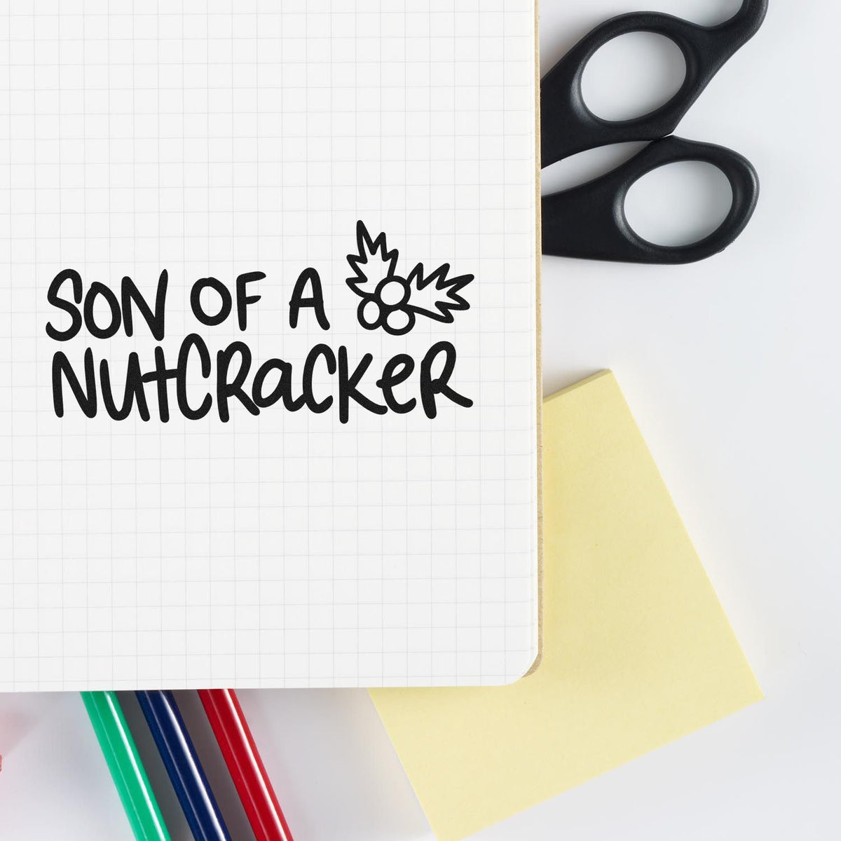 Black custom stamp-style "Son of a Nutcracker" with holly seal on grid notebook, beside black scissors, yellow note, and pens.