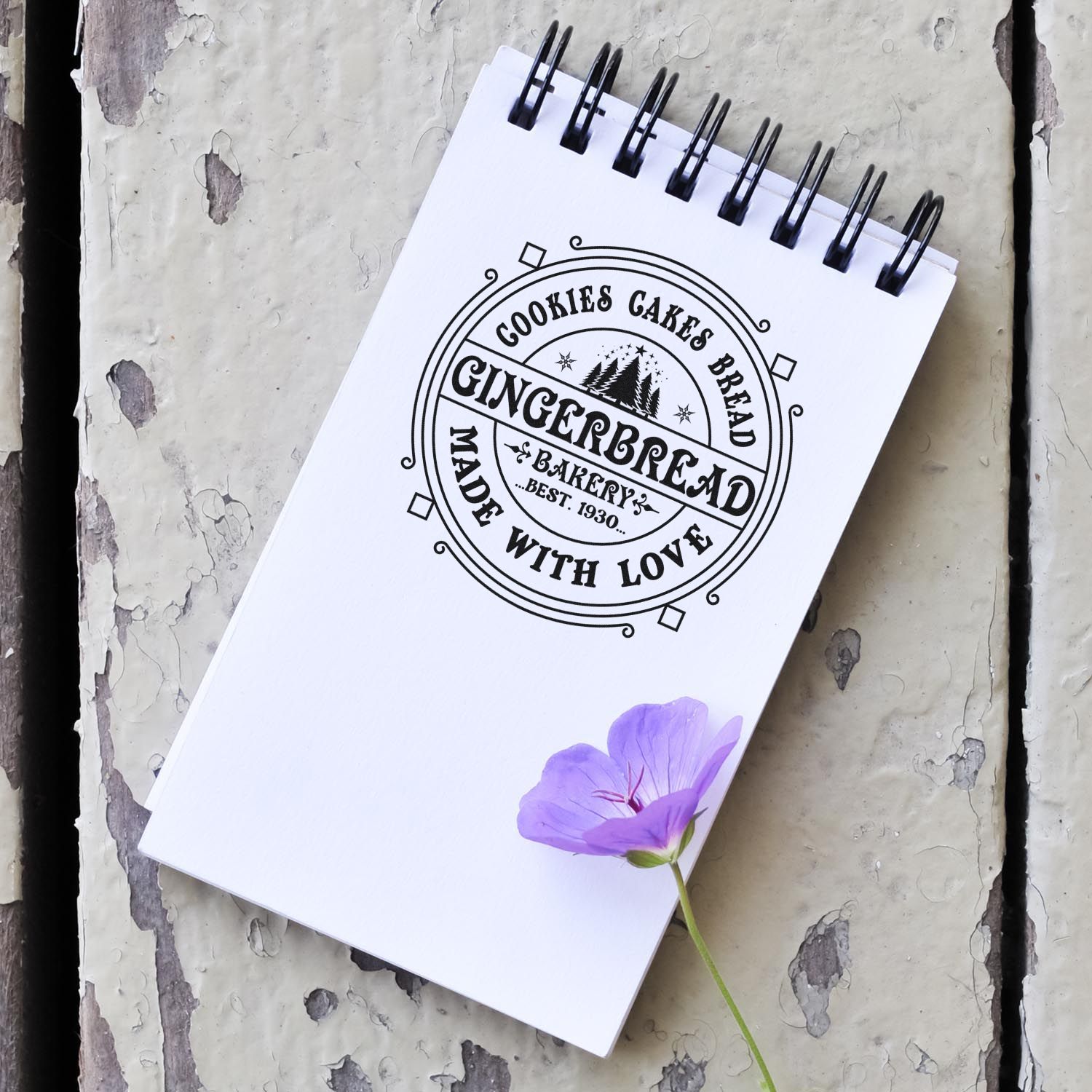 White spiral notebook featuring a black custom bakery stamp seal reading Gingerbread Made with Love, with a purple flower.