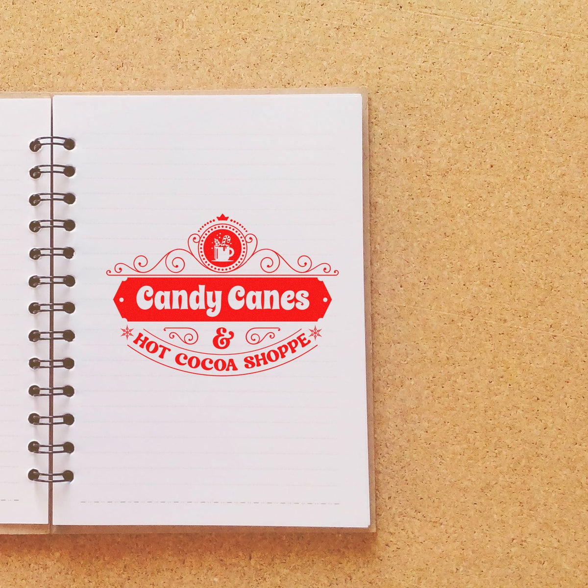 Red decorative stamp seal for Candy Canes & Hot Cocoa Shoppe on a white spiral notebook over cork background.