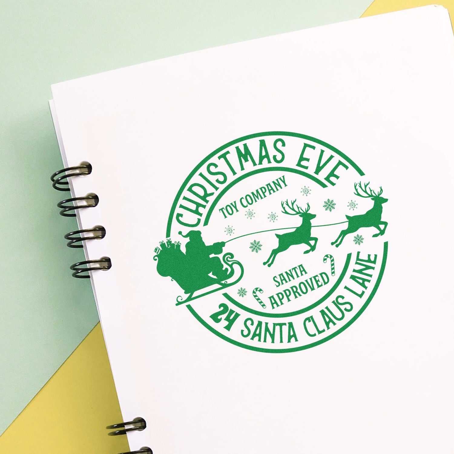 Green Christmas Eve stamp seal on white paper, with Santa sleigh, reindeer, snowflakes, and holiday text in spiral notebook.
