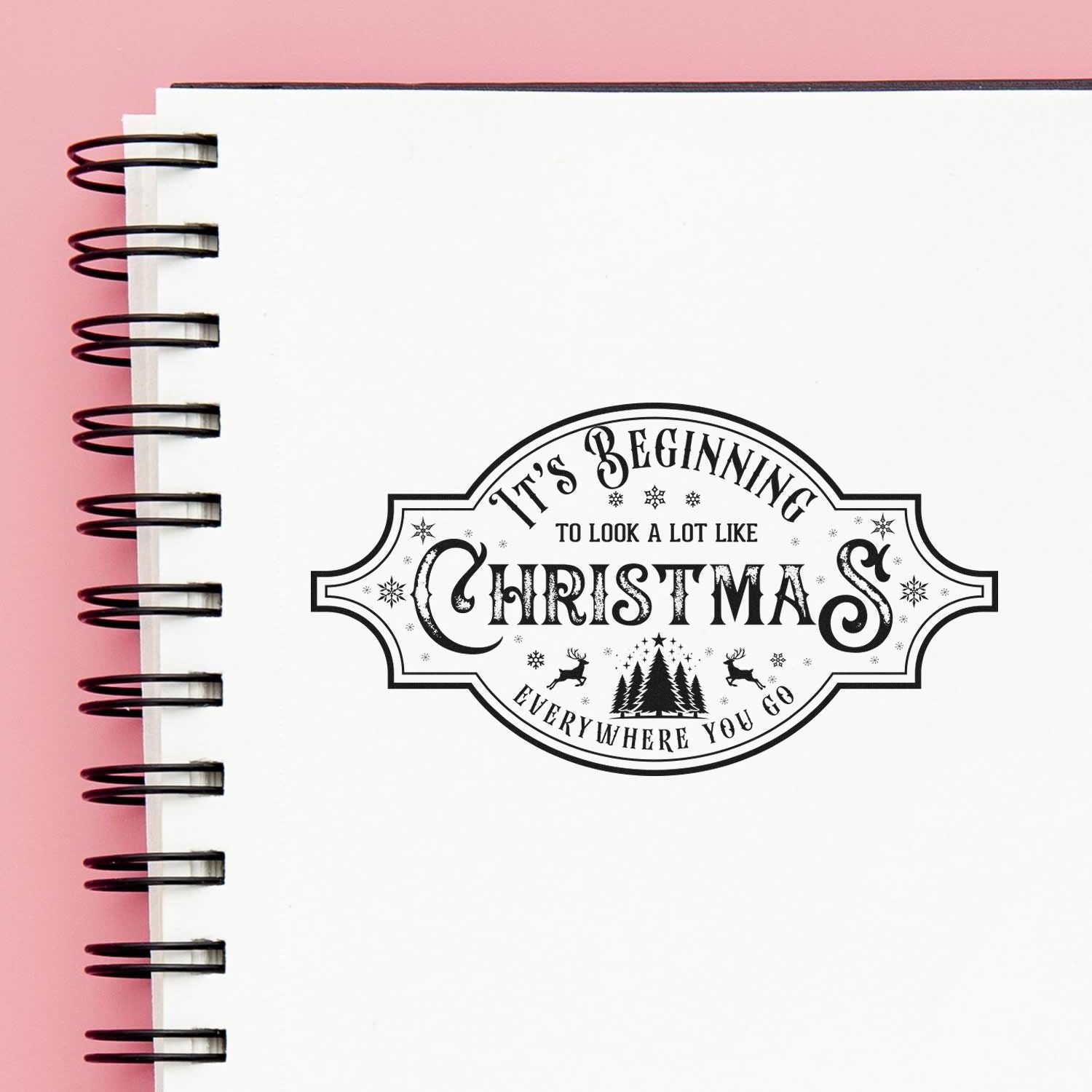 Black Christmas-themed stamp seal on white spiral notebook, with vintage lettering and pink background.