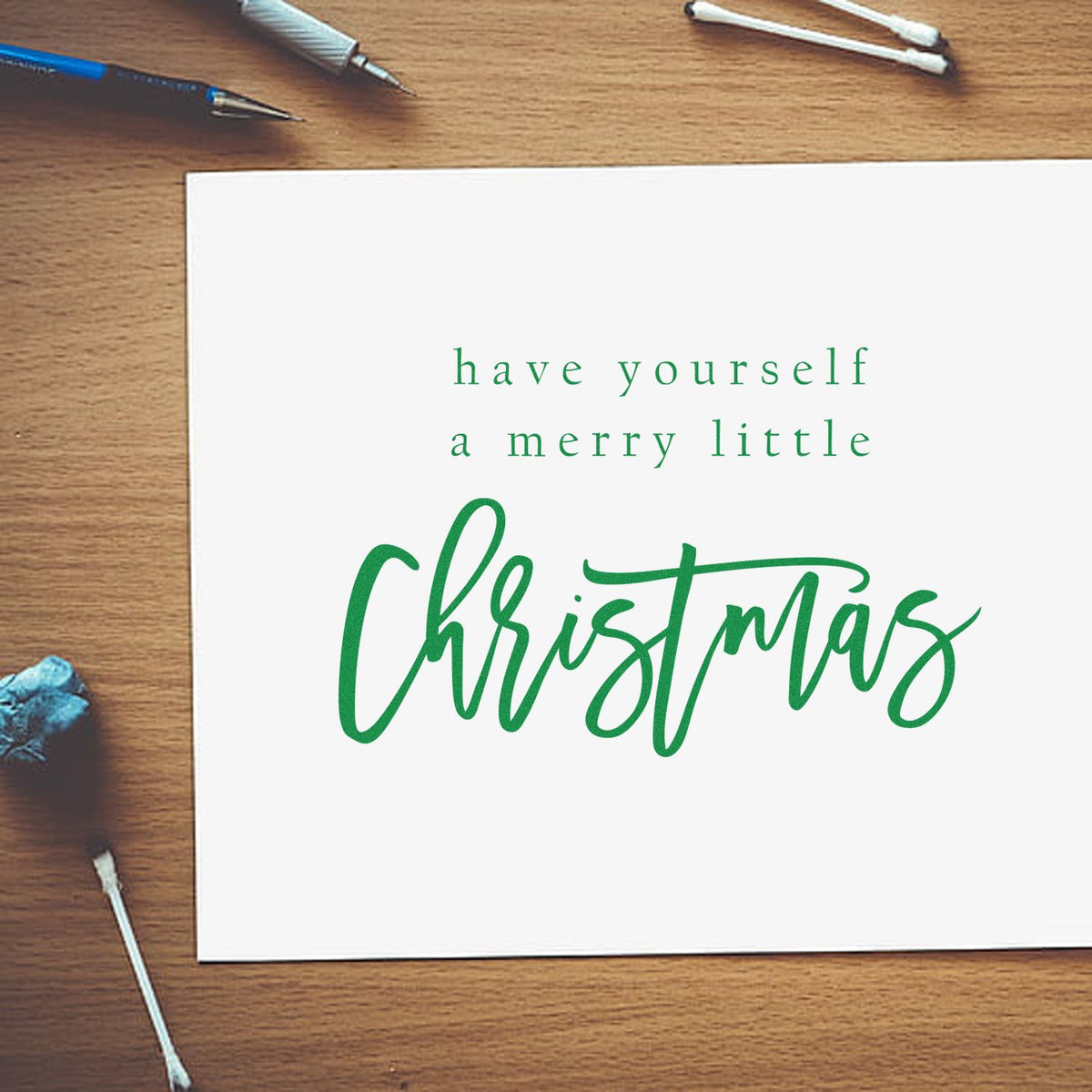 Green Christmas stamp and seal design on a white card, with have yourself a merry little Christmas in green on a wood desk.