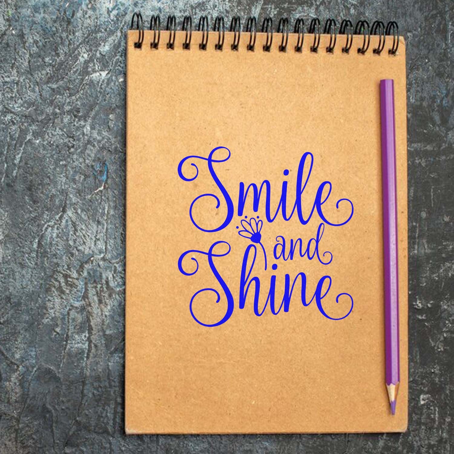 Kraft spiral notebook featuring a blue Smile and Shine stamp/seal design with a purple pencil on a dark textured background.