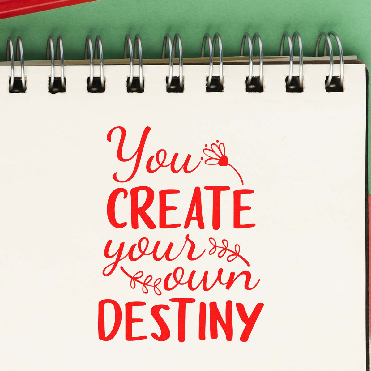 Red seal-style slogan stamp on white spiral notebook reads You create your own destiny with floral accents.