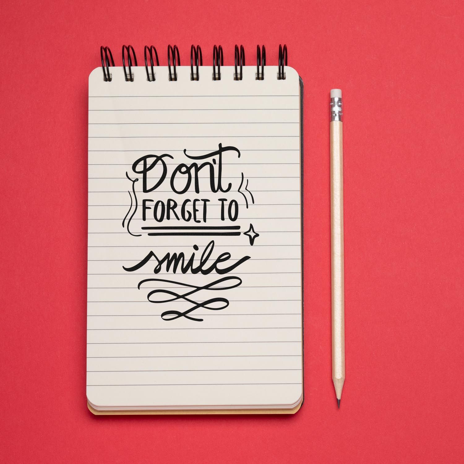 Black script stamp design reading Dont forget to smile on a lined notebook, beside a pencil on a red background.