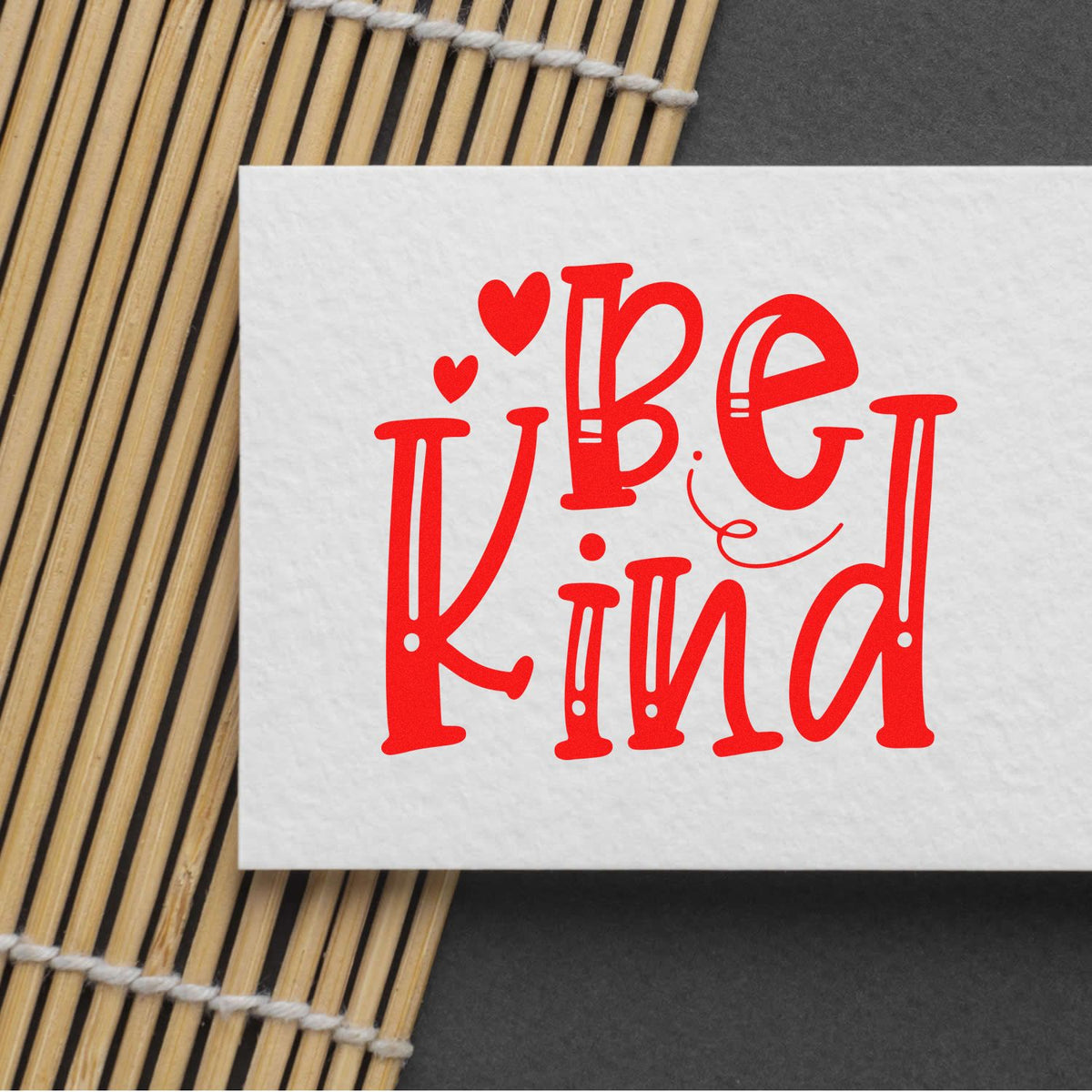 Red Be Kind stamp and seal design with hearts on white paper, set on a bamboo mat against a dark background.