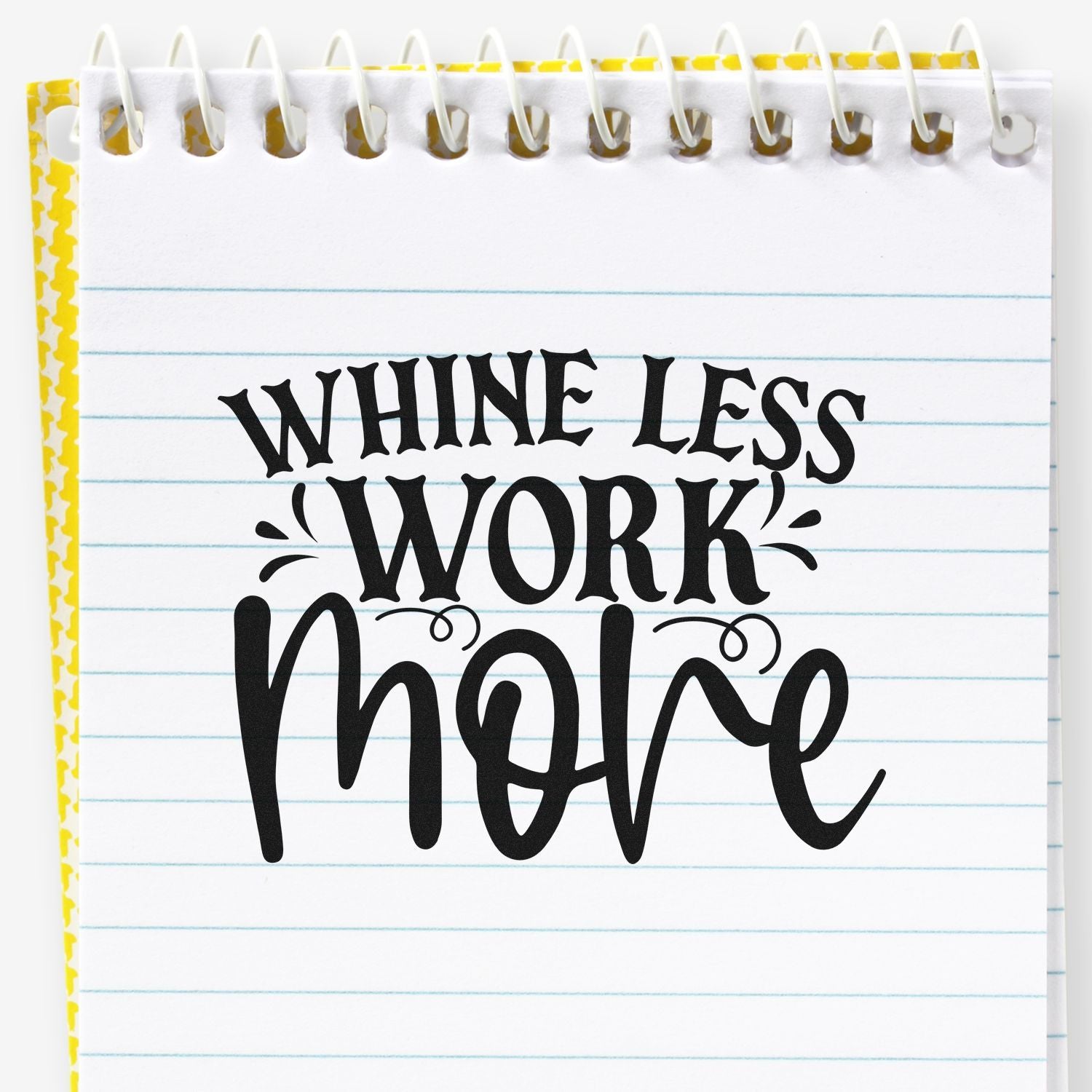 Black decorative stamp on white spiral notebook reads Whine Less Work More in bold lettering, with blue lines and yellow border.