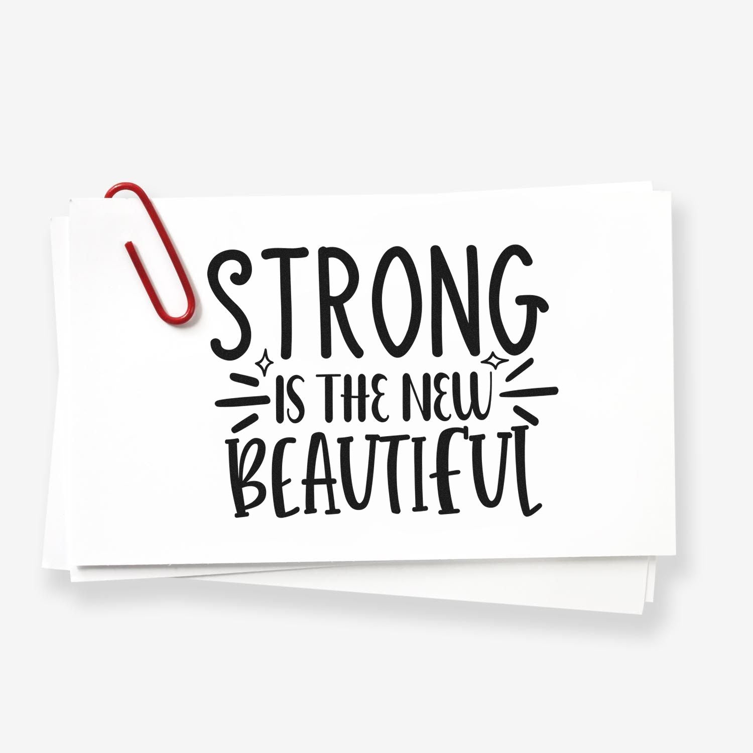 Black custom stamp/seal design reading Strong Is the New Beautiful on white paper, accented by a red paperclip.