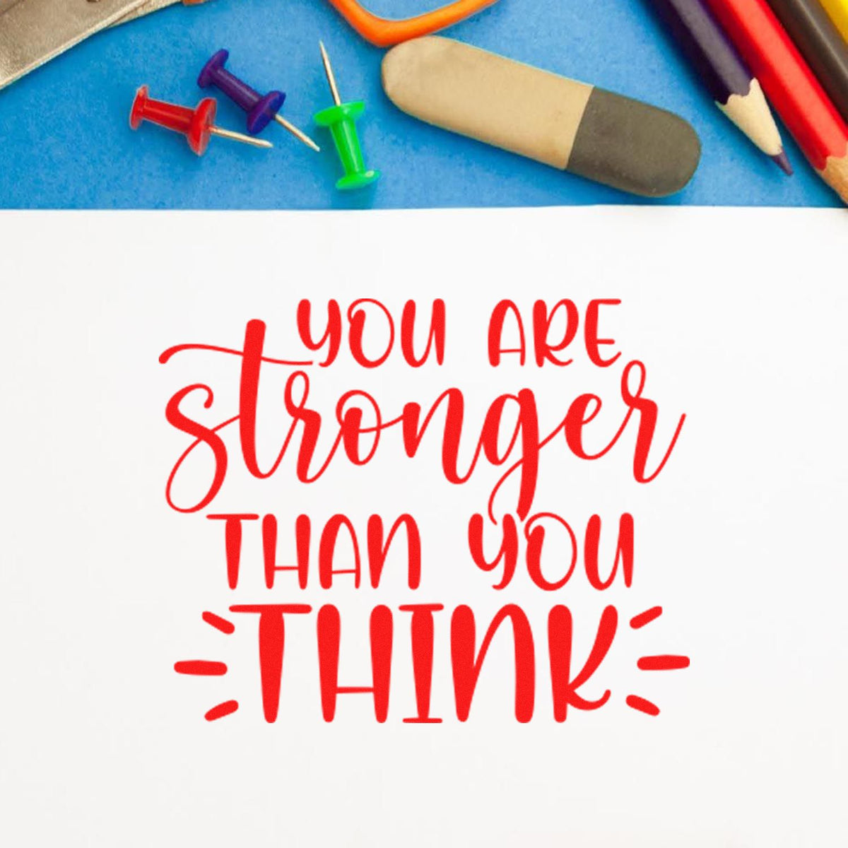 Red motivational rubber stamp reading "You are stronger than you think" on white paper, with pencils, push pins, and eraser on blue.