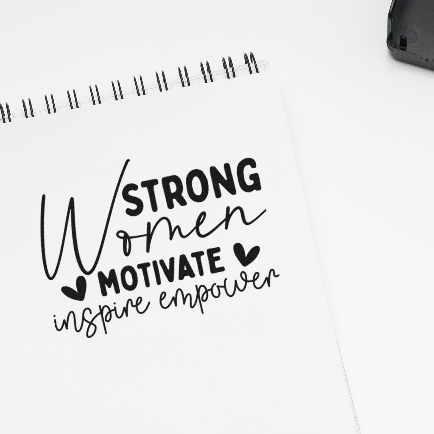 Black stamp-and-seal design on white spiral notebook, reading Strong Women Motivate Inspire Empower with hearts.