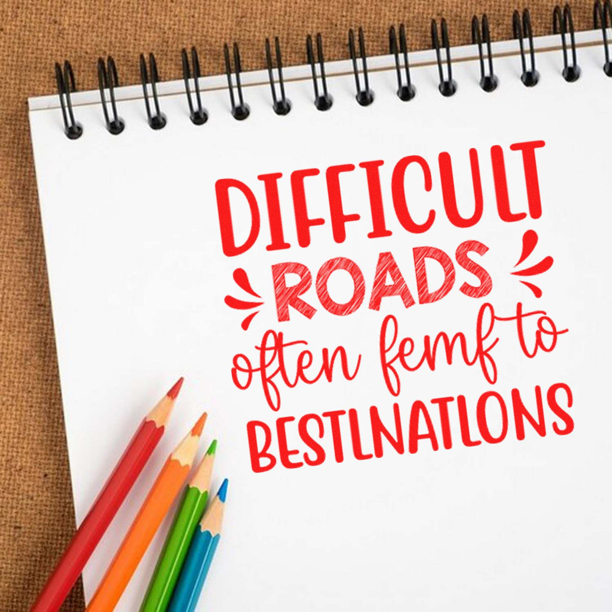 Red stamp-style motivational text on white spiral notebook with colored pencils and burlap background.
