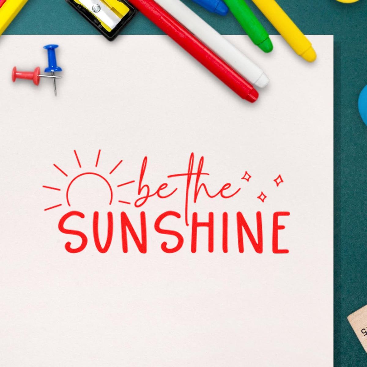 Red stamp and seal reading be the SUNSHINE with sun doodle on white paper, framed by colorful pens and pins.
