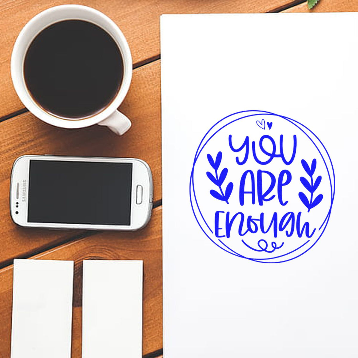 Blue You Are Enough circular stamp seal on white paper beside a coffee cup and smartphone on a wooden desk.