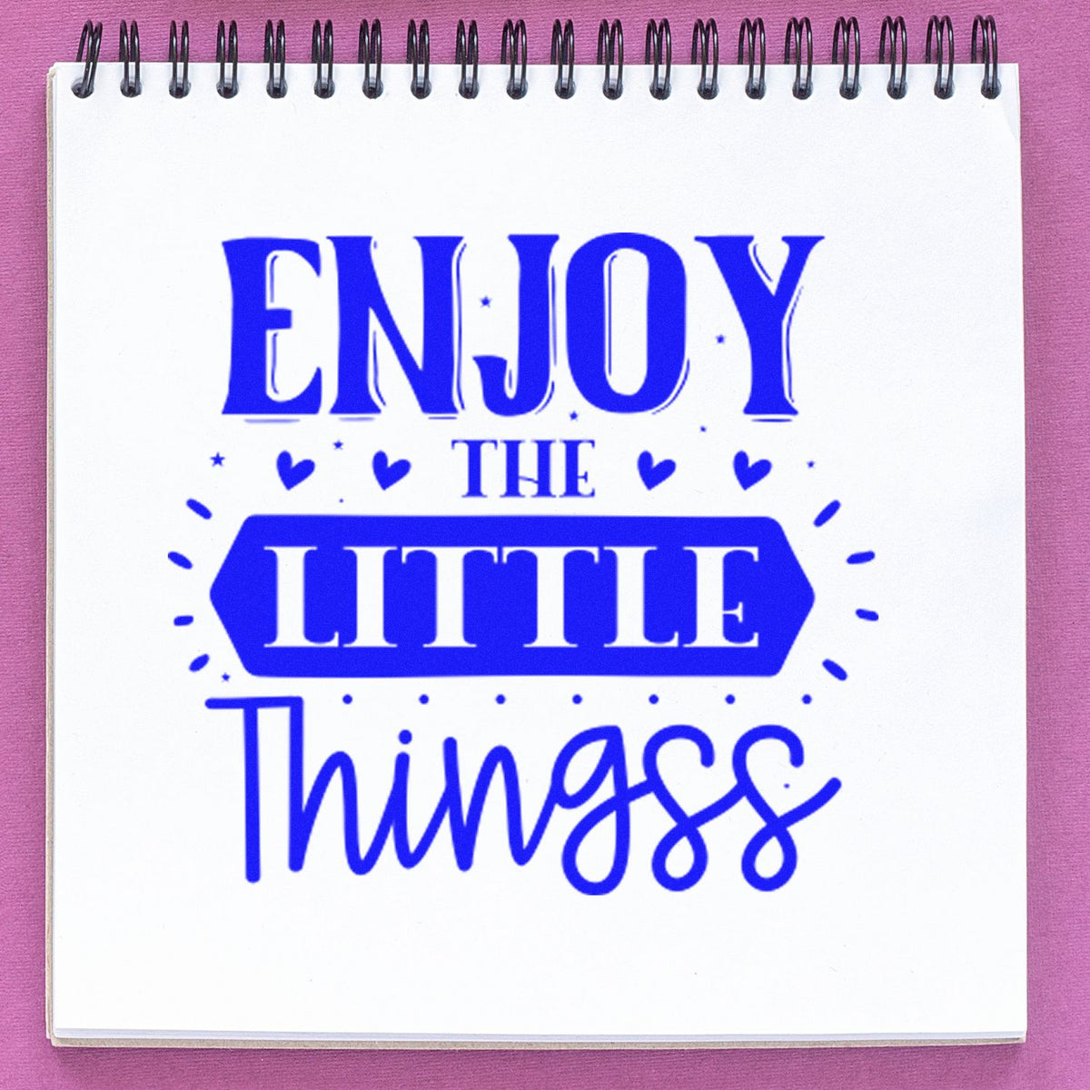 Blue decorative stamp/seal design with Enjoy the Little Things in bold blue on a white spiral notebook, accented by hearts and stars on pink.