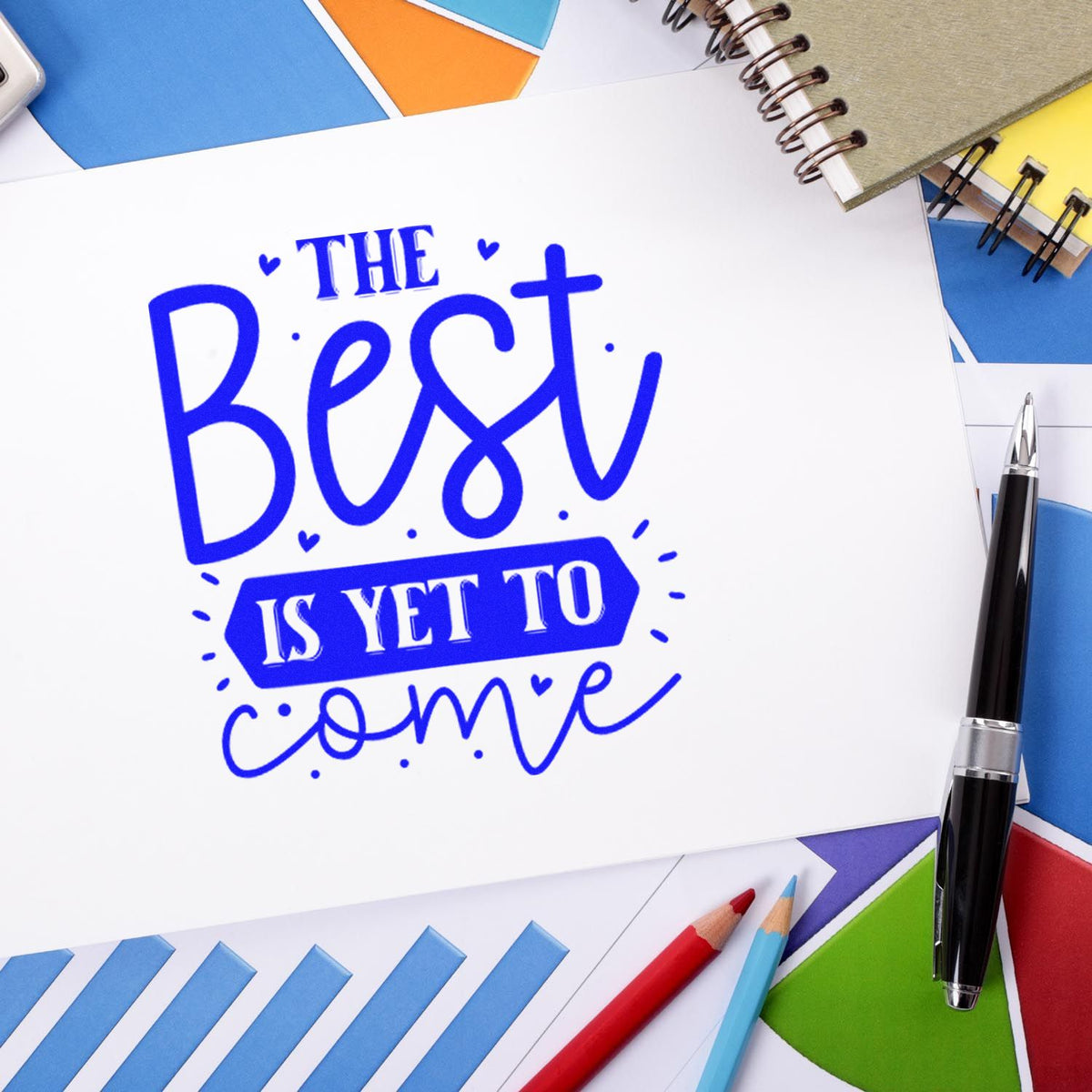 Blue custom stamp and seal reading The Best Is Yet to Come, on white paper with colorful notebooks, pens, and pencils.