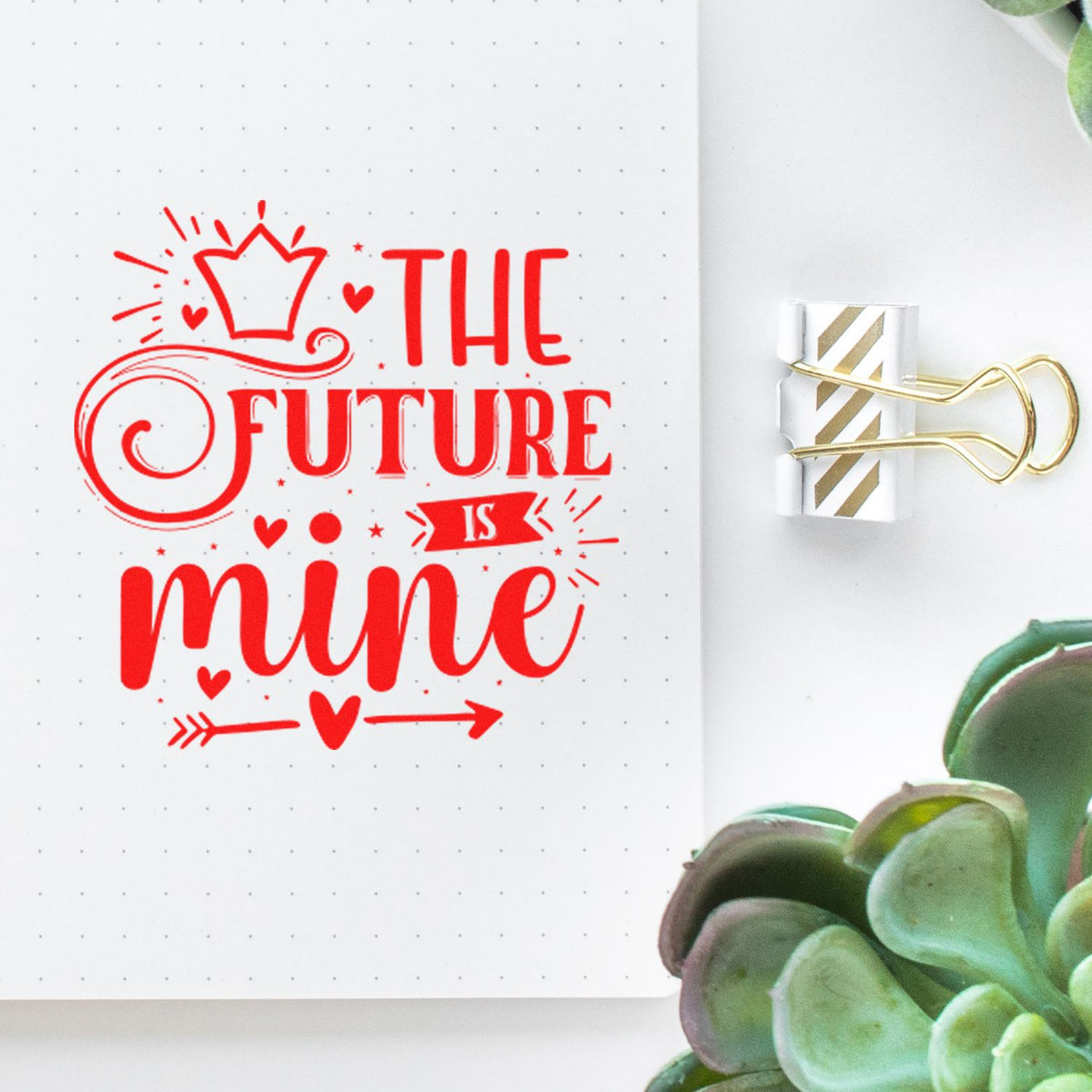 Red decorative stamp reading The Future Is Mine on white dotted paper, clipped with gold striped binder clip, beside green succulent.