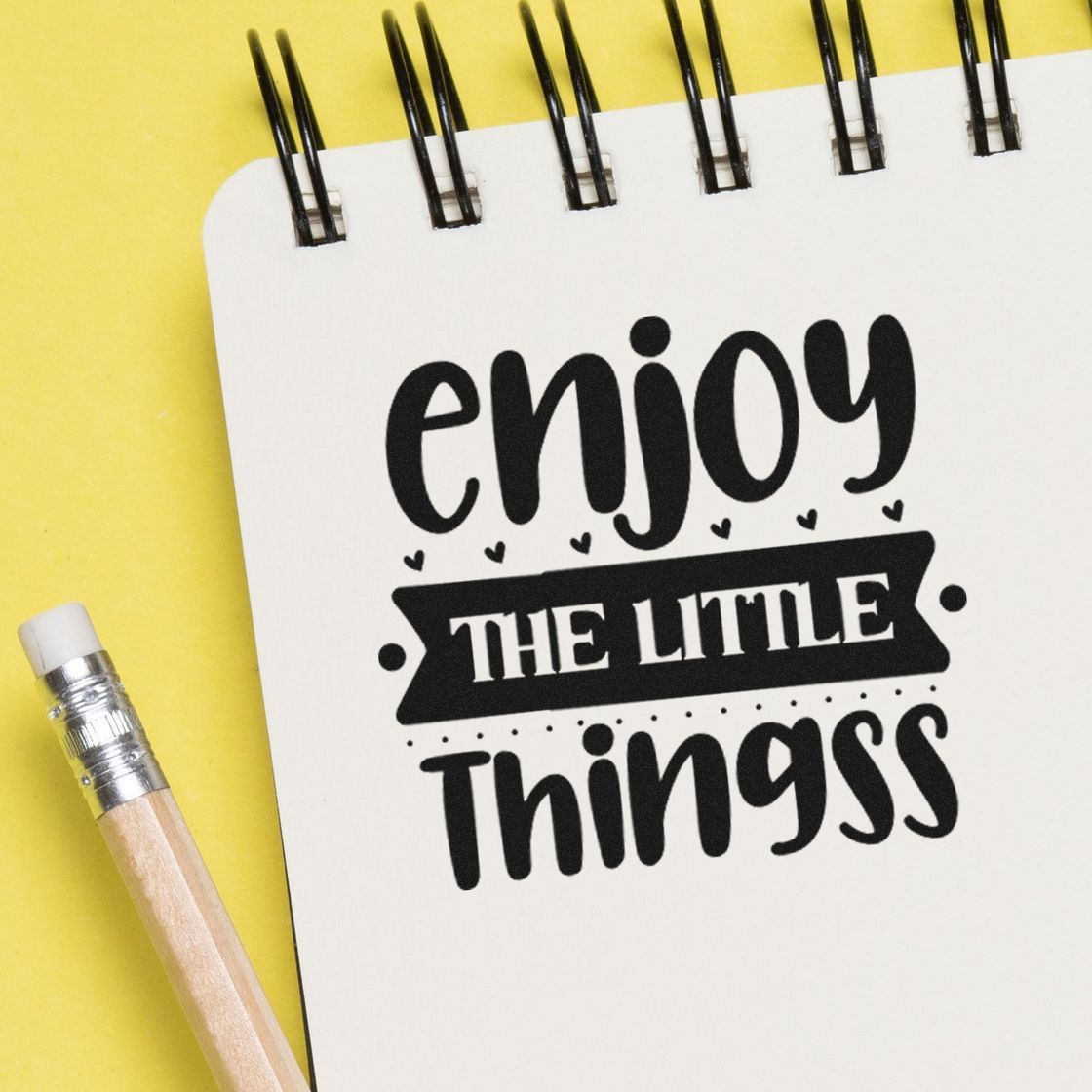 Black stamp and seal design on white spiral notebook reading enjoy the little things, with yellow background and pencil.