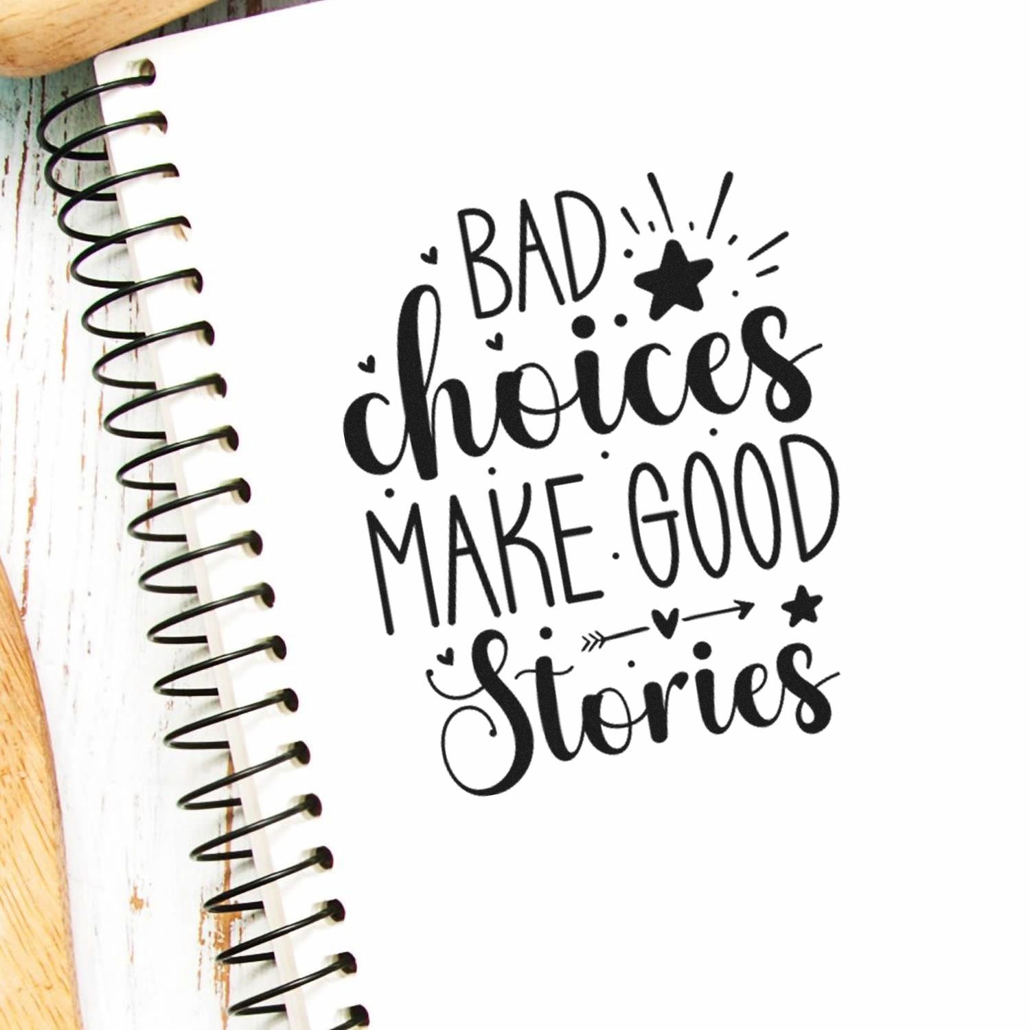 Black decorative stamp and seal reading Bad Choices Make Good Stories on a white spiral notebook page.