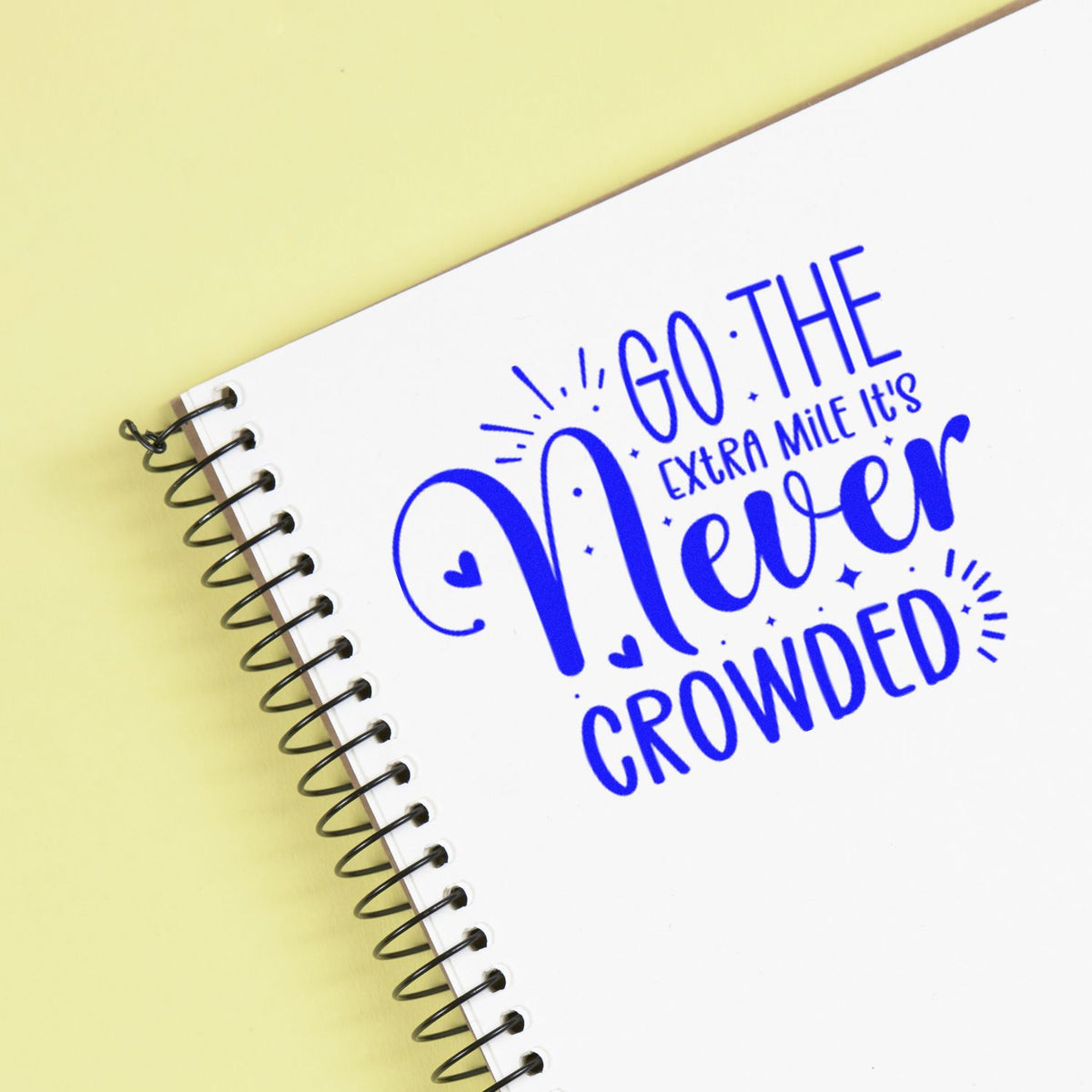 Blue motivational stamp seal on a white spiral notebook over pale yellow background, reading Go the extra mile, its never crowded.