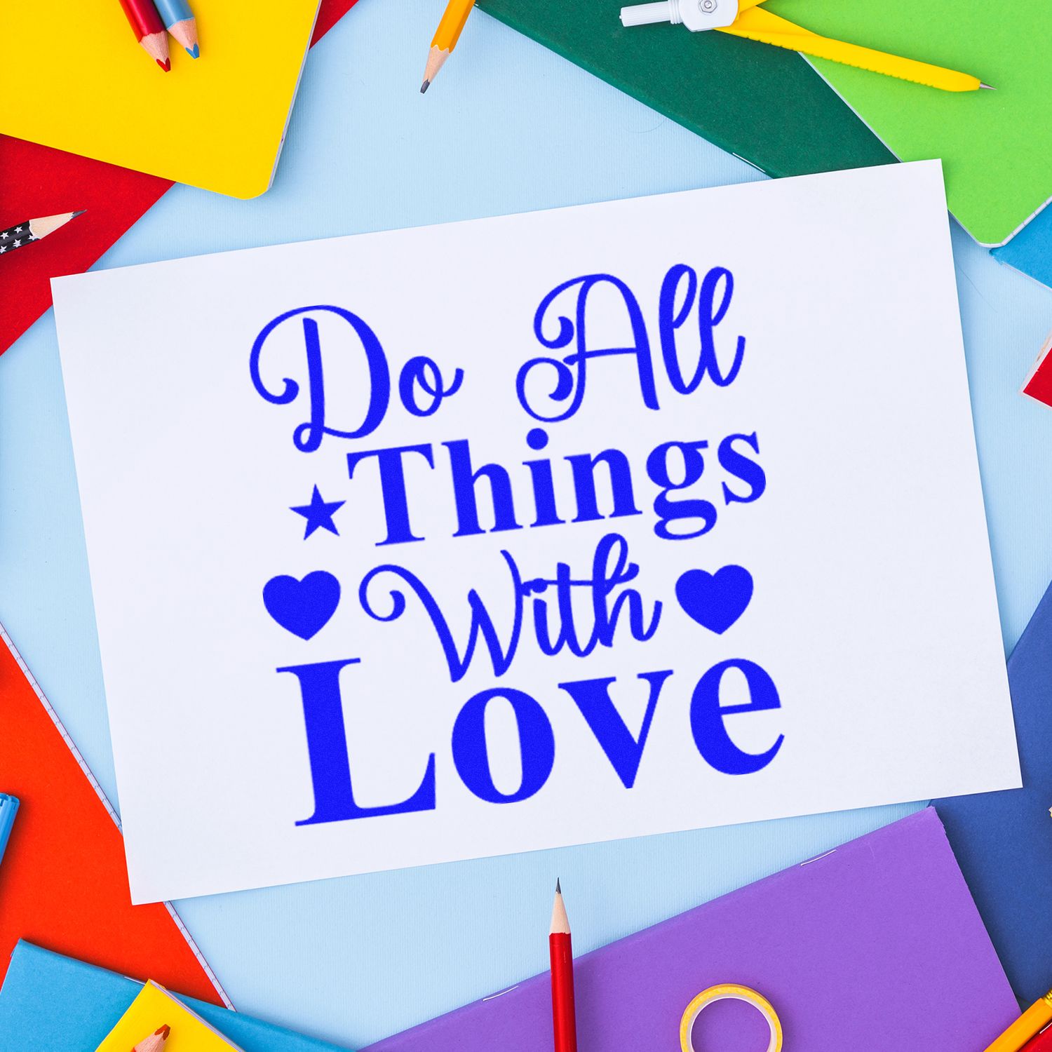 Blue Do All Things With Love stamp and seal on white card, framed by colorful notebooks and pencils.