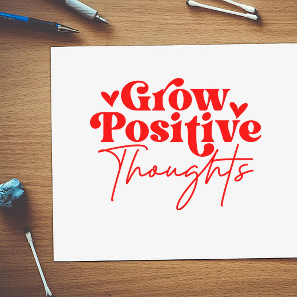 Red custom stamp and seal reading Grow Positive Thoughts on white paper, with hearts on a wood desk.