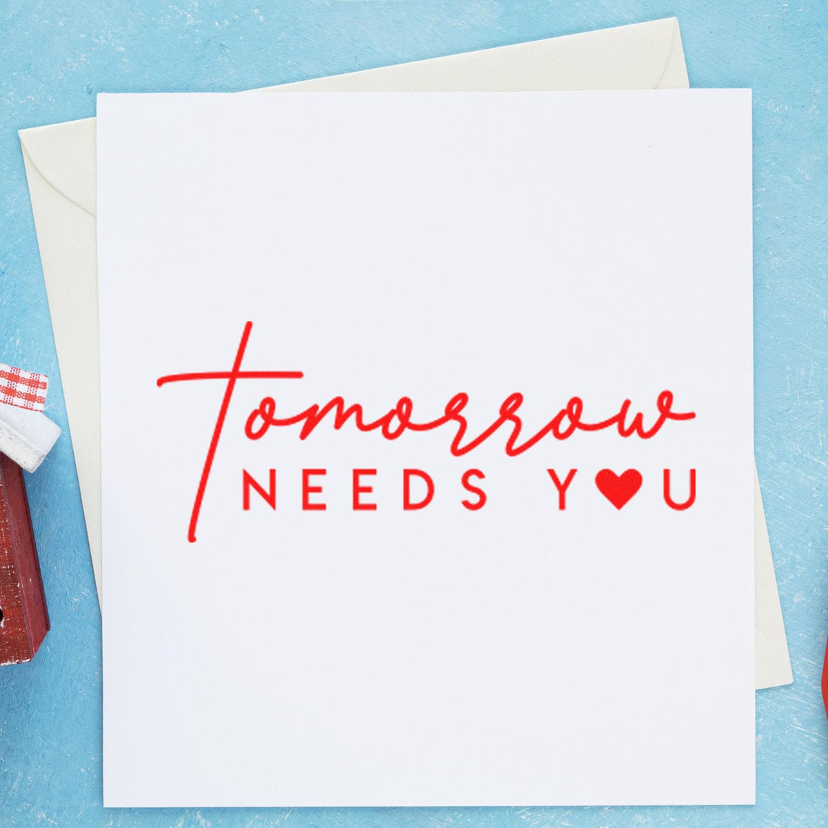 Red stamp and seal impression on white card reading Tomorrow needs you with heart icon, over a blue background.